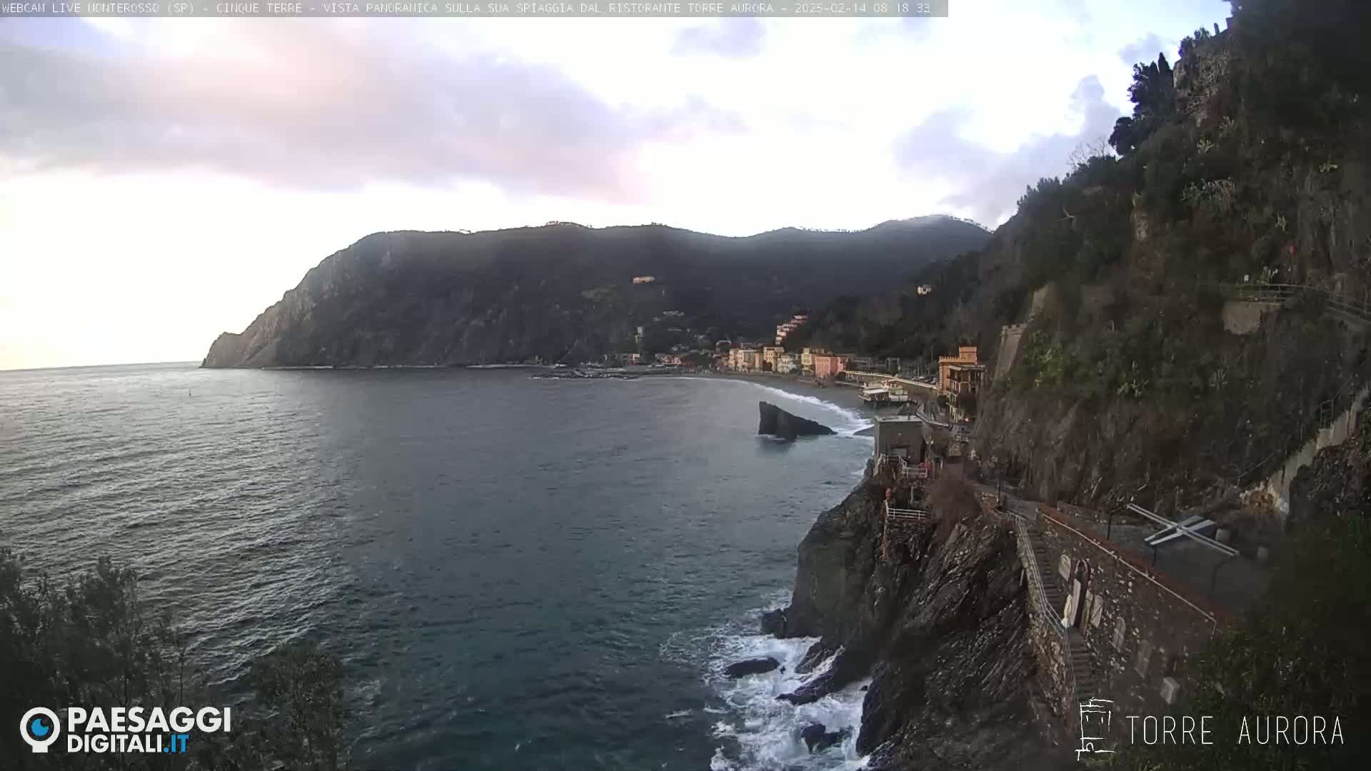 A coastal town nestled between a rocky cliff and the sea under an overcast sky, with waves crashing against the shore.