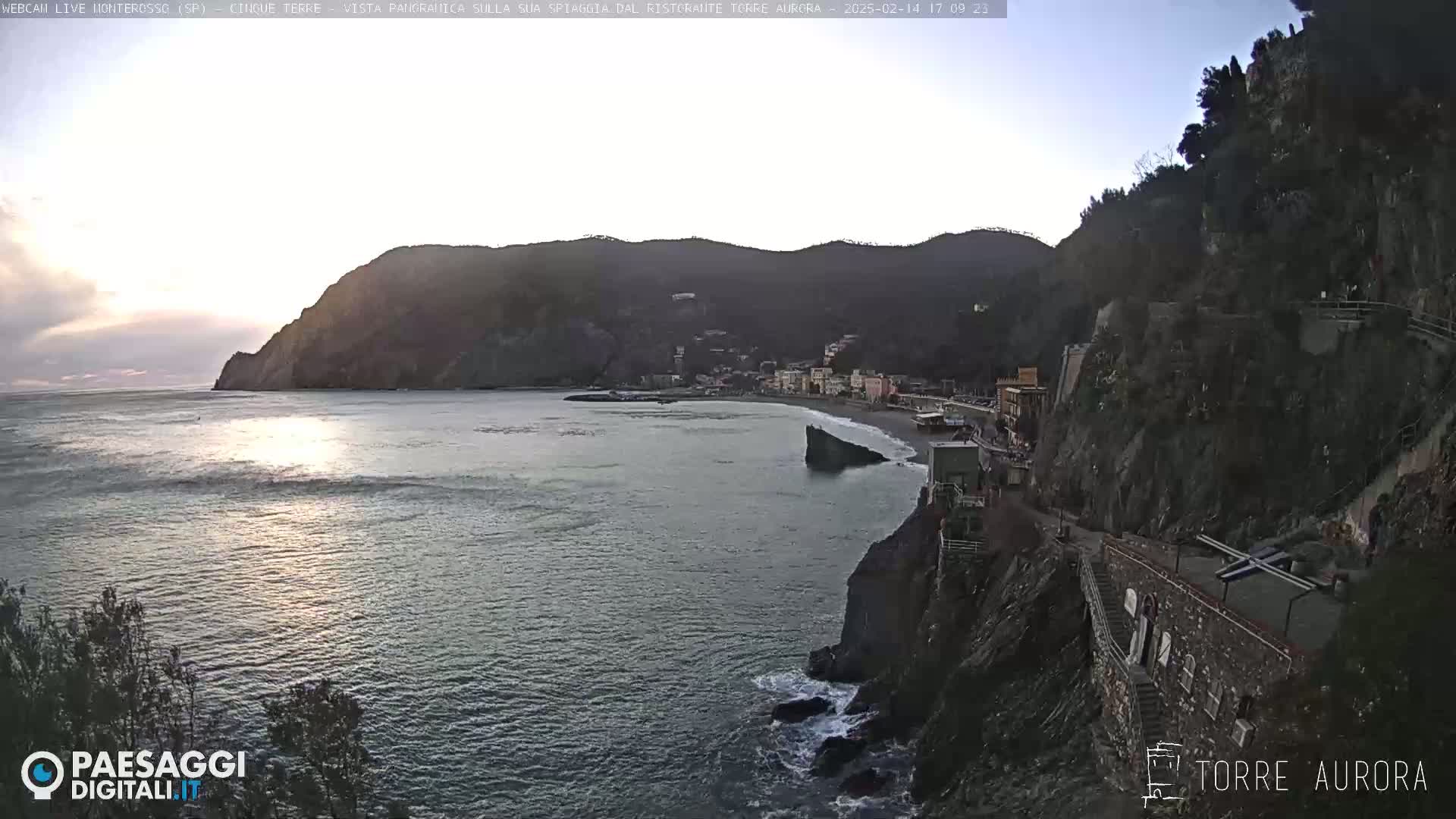 A coastal town nestled between dark, rocky cliffs, with a calm sea reflecting the setting sun under a mostly clear sky.