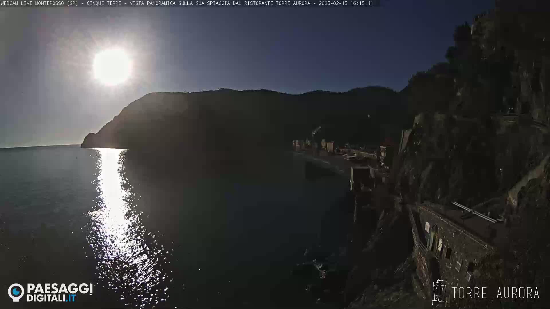 A bright sun shines over a calm sea, reflecting on the water near a rocky shoreline with buildings on the cliffside.