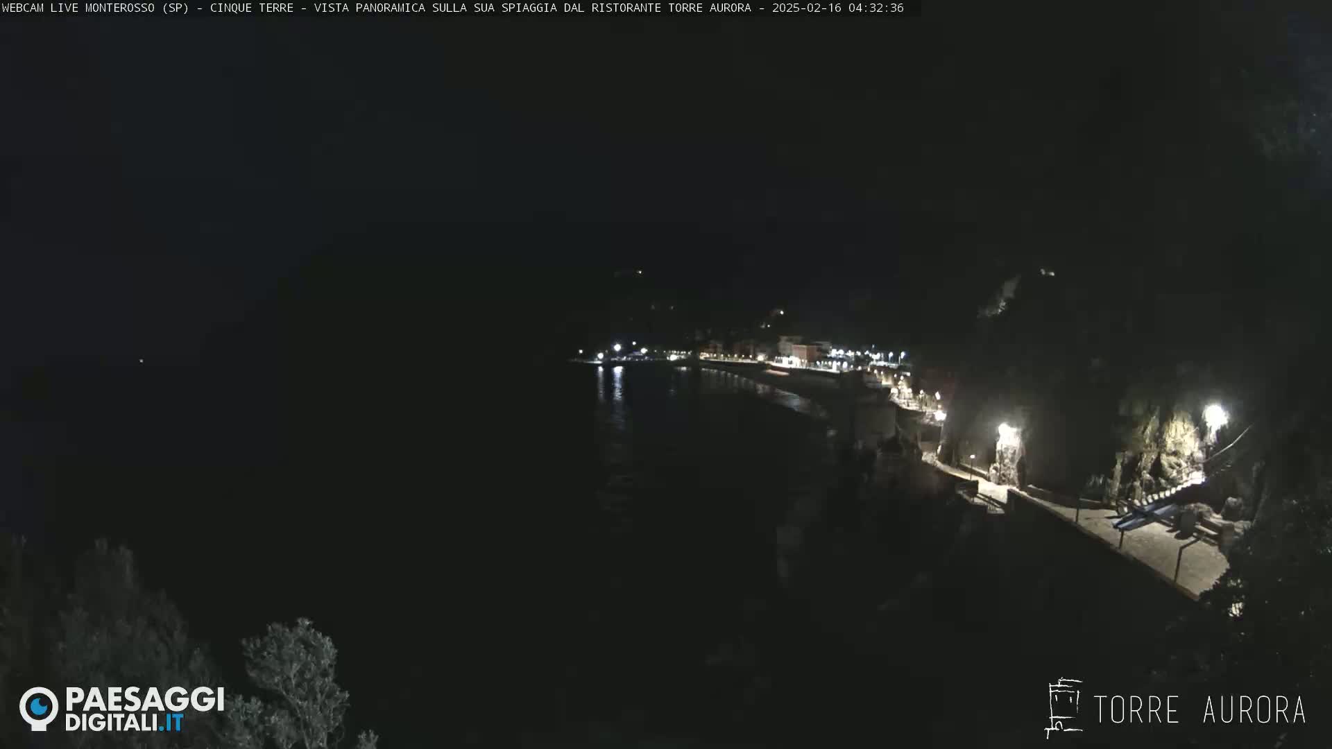 A nighttime, long shot of a dark coastline with a few lights visible along a paved walkway and buildings, overlooking a calm body of dark water.