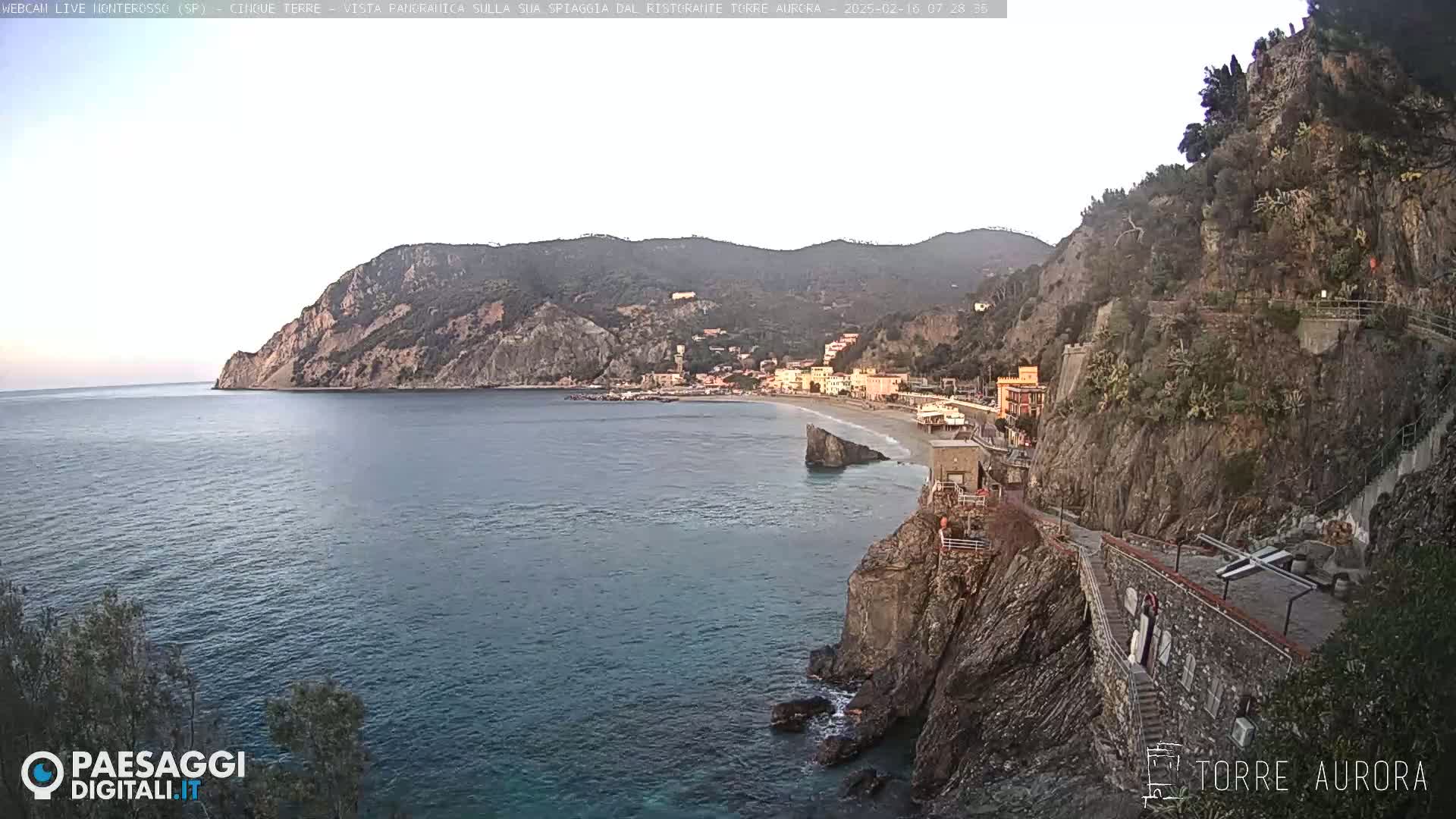 A coastal town nestled between a rocky cliff and the calm sea under a clear, light sky.