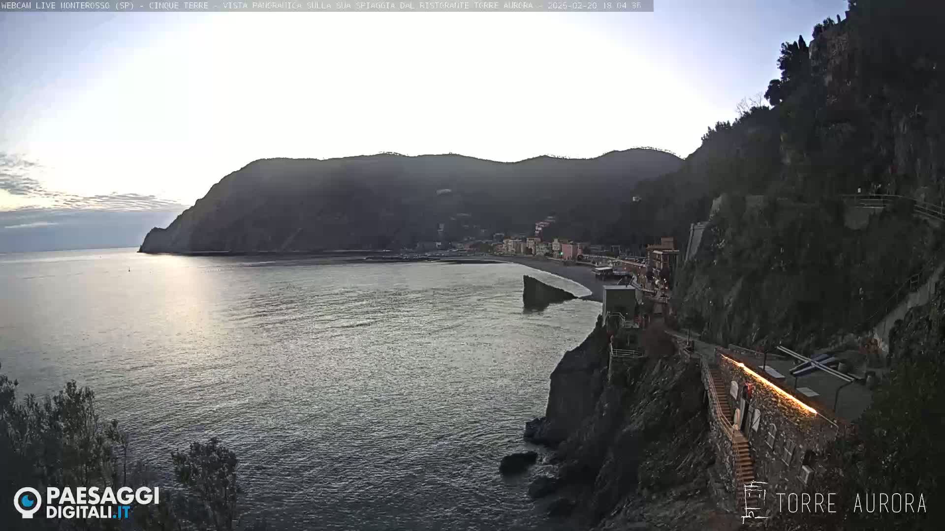 A coastal town nestled between dark, rocky cliffs and a calm, gray-blue sea under a dusky sky.