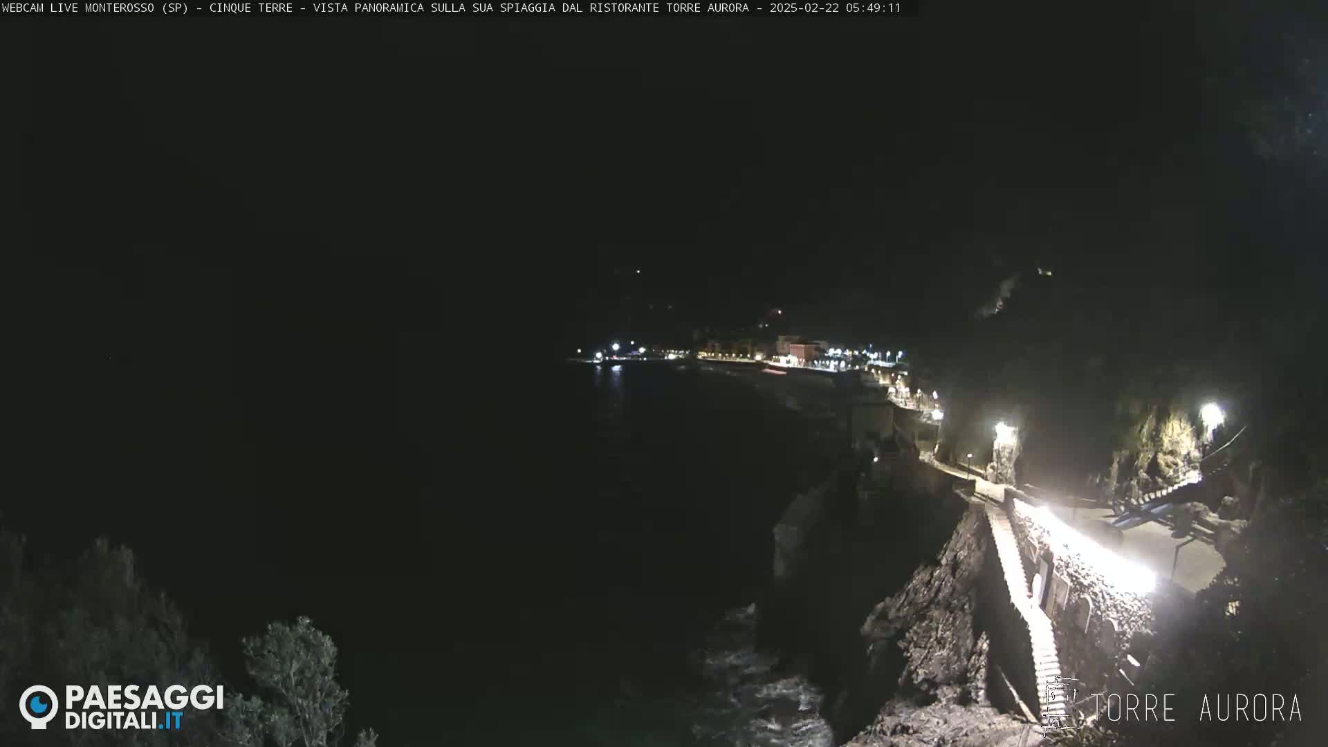 A nighttime, panoramic view of a coastal town with a stone staircase descending to a dark, wavy ocean under a clear, starless sky.