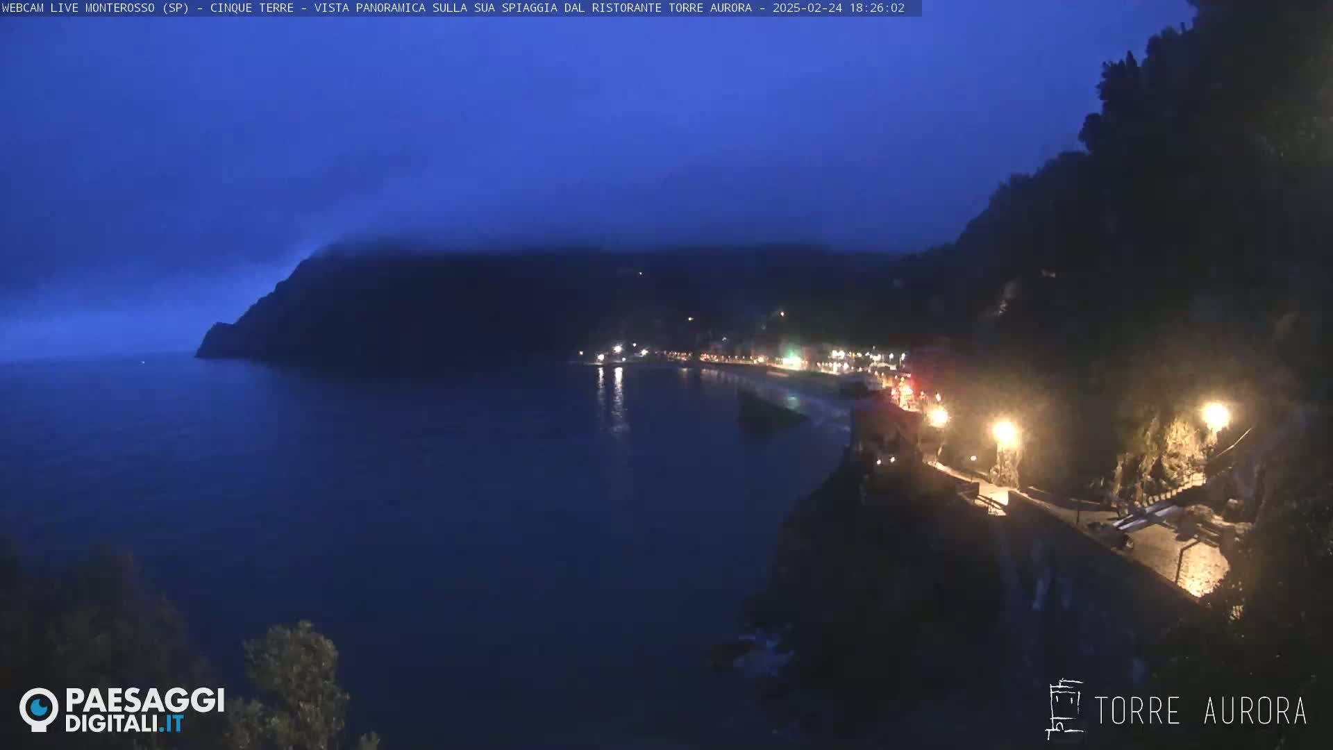 A nighttime coastal scene shows a dark, hazy sky over a calm, dark blue sea, with a dimly lit town nestled along a cliffside.