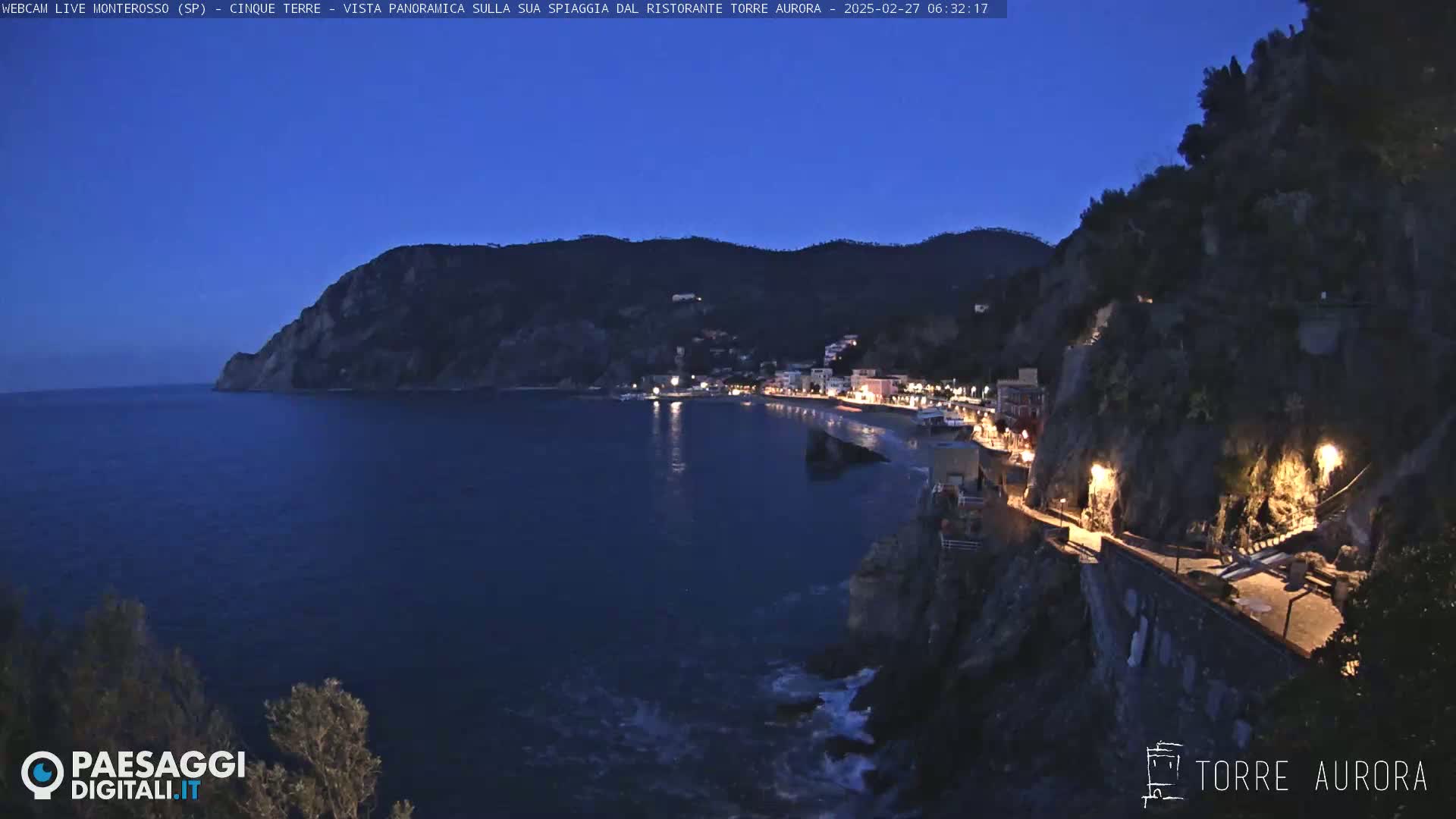 A coastal town at night with lights illuminating the buildings and a pathway along the cliffside, under a dark blue, twilight sky.