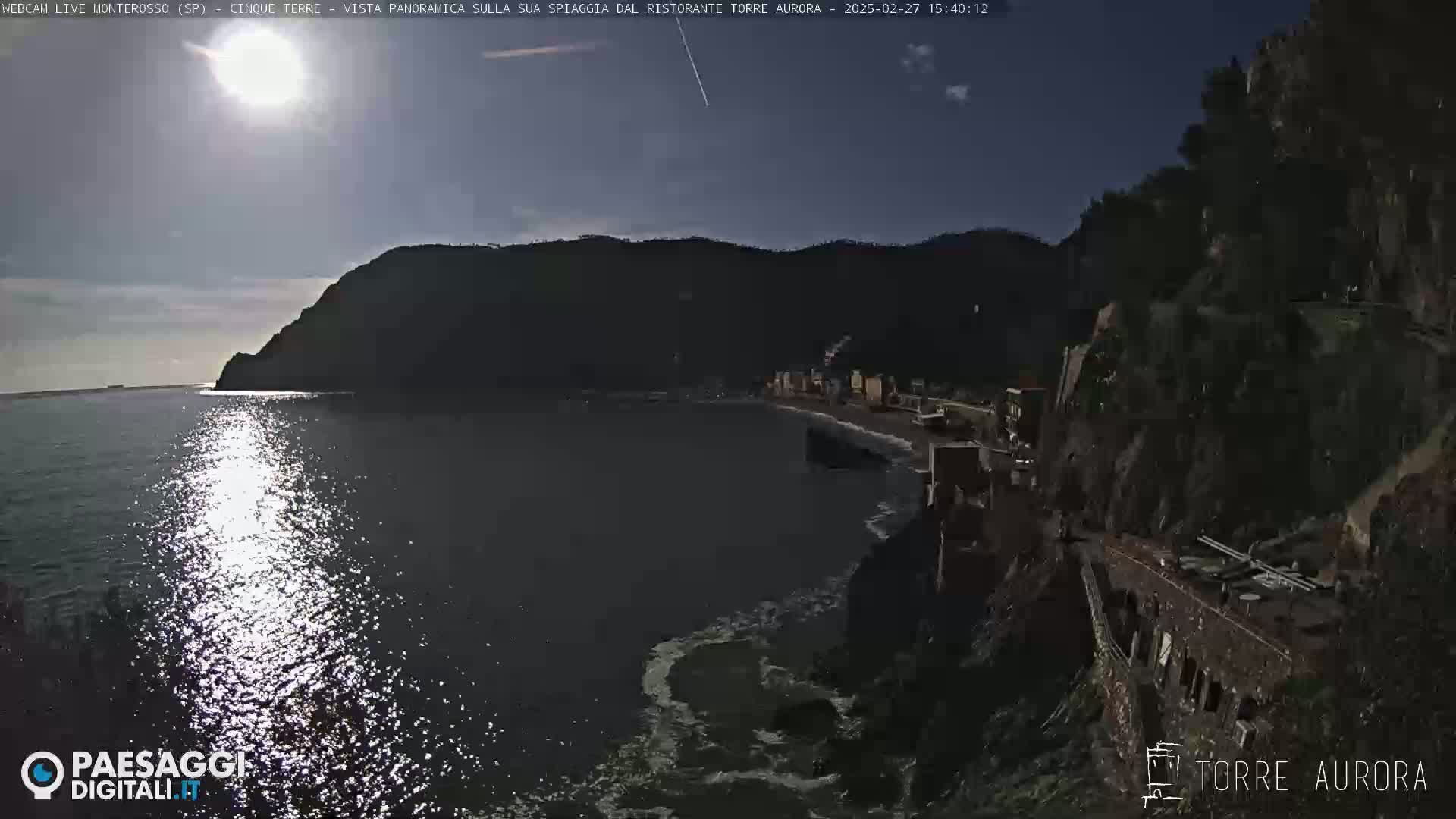 A daytime coastal scene shows a calm sea reflecting sunlight, with a dark rocky shoreline and hills in the background under a mostly clear sky.