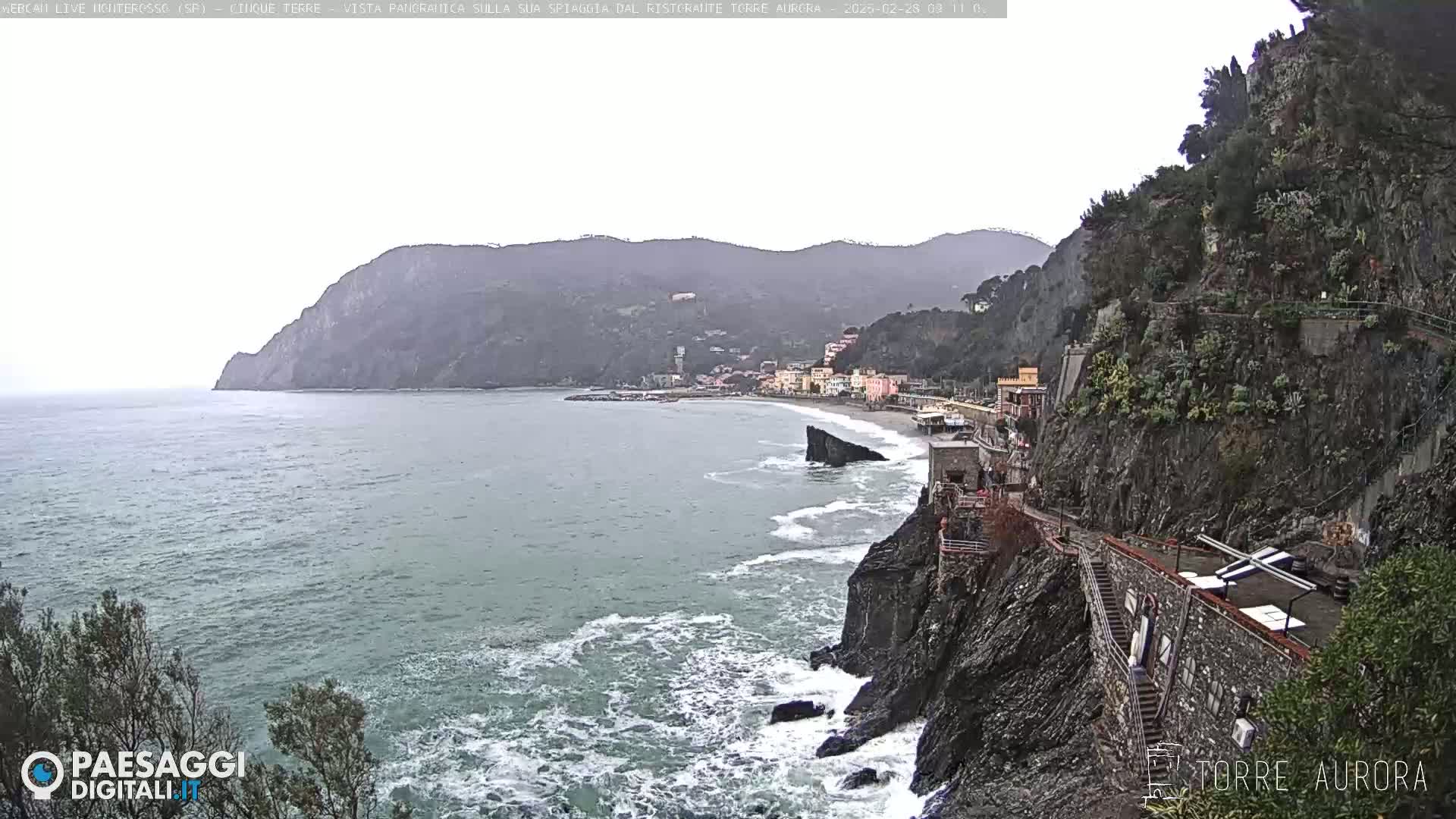 A coastal town nestled between steep cliffs is seen on an overcast day, with waves crashing against the rocky shore.