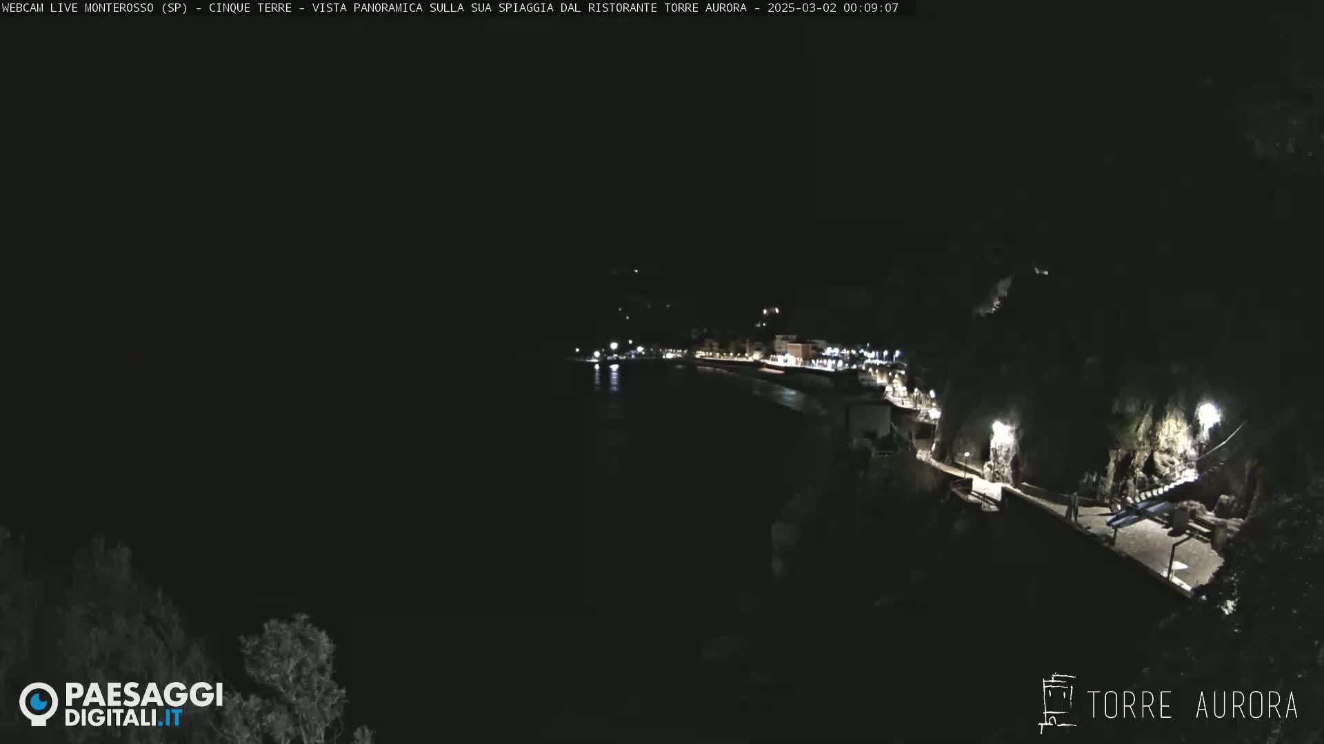 A nighttime, coastal view shows a dark body of water next to a dimly lit shoreline with buildings and a pathway.