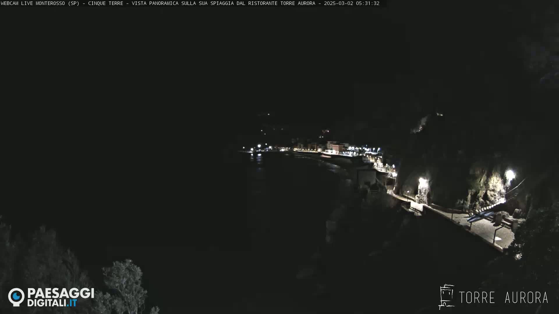 A nighttime, coastal town scene shows a dark shoreline with sparsely lit buildings and a walkway along a cliffside.