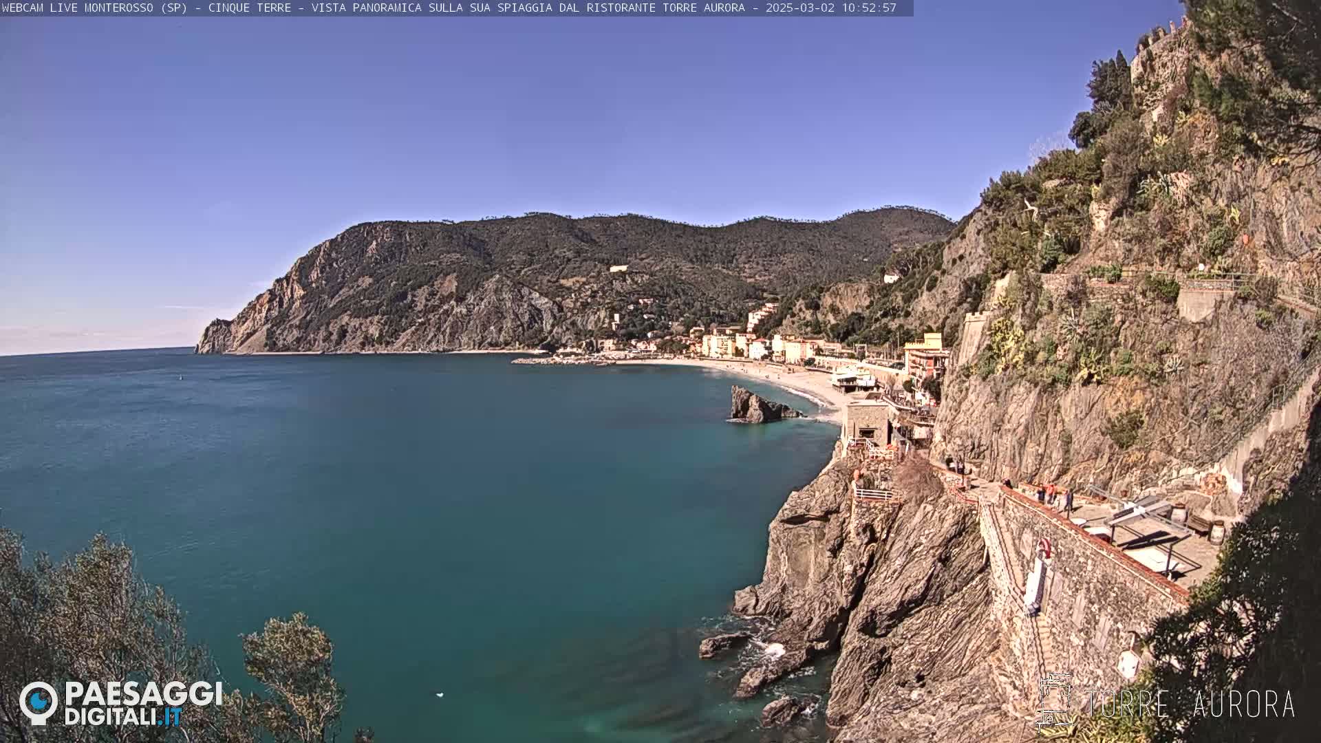 A coastal town nestled between rocky cliffs and a calm, teal sea under a clear, sunny sky.