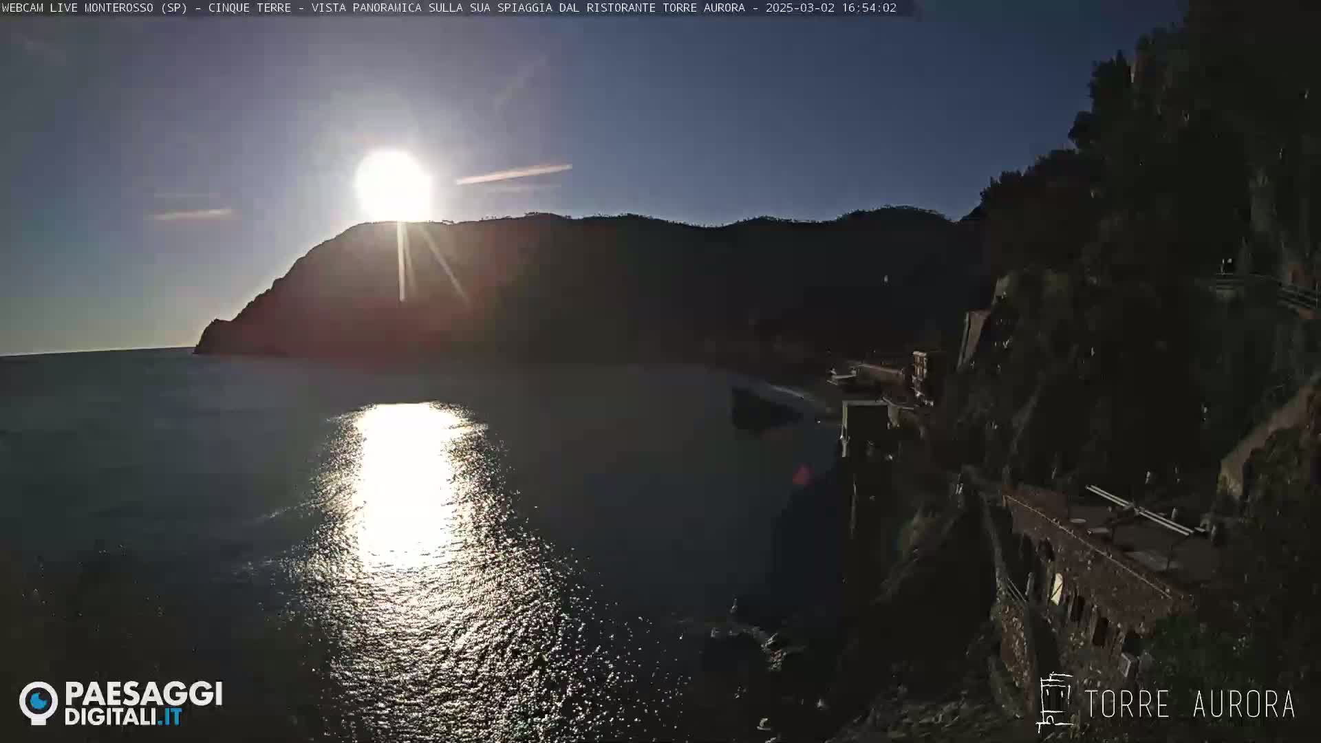 A bright sun shines over a calm body of water, reflecting its light, with dark, rocky hillsides on either side.