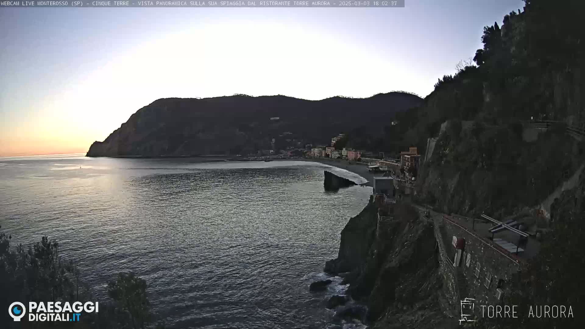 A coastal town nestled between dark hills overlooking a calm sea under a twilight sky.
