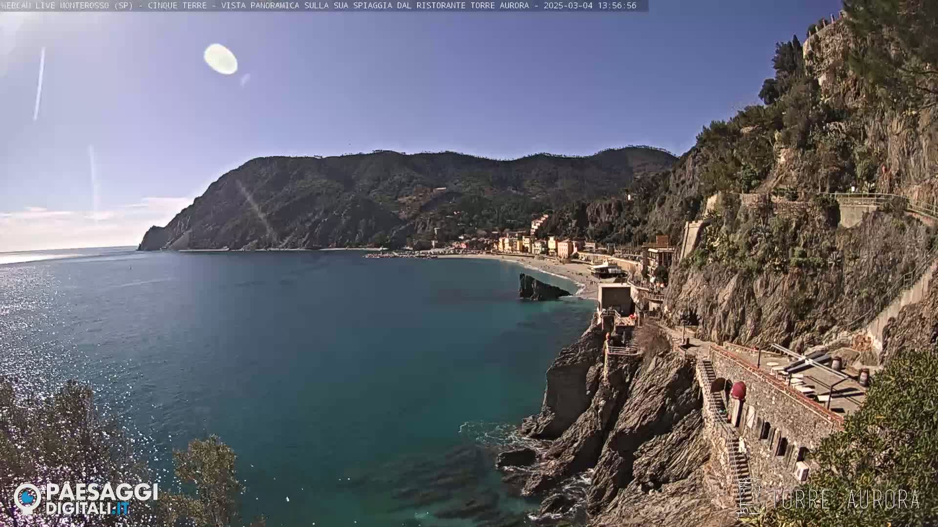A sunny day overlooking a coastal town nestled between a teal sea and steep, rocky cliffs, with a pathway winding along the cliffside.