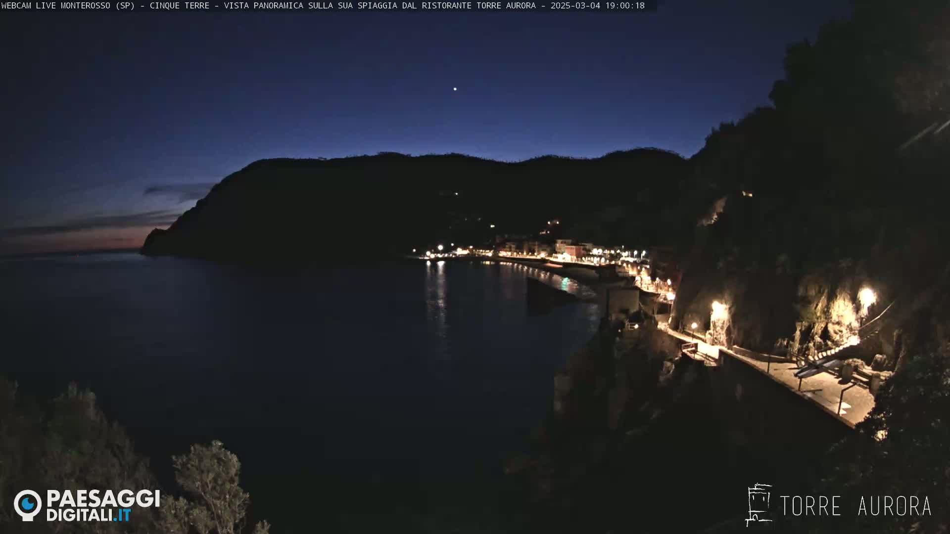 A nighttime coastal town with lights illuminating a walkway along the water's edge under a dark blue, twilight sky.