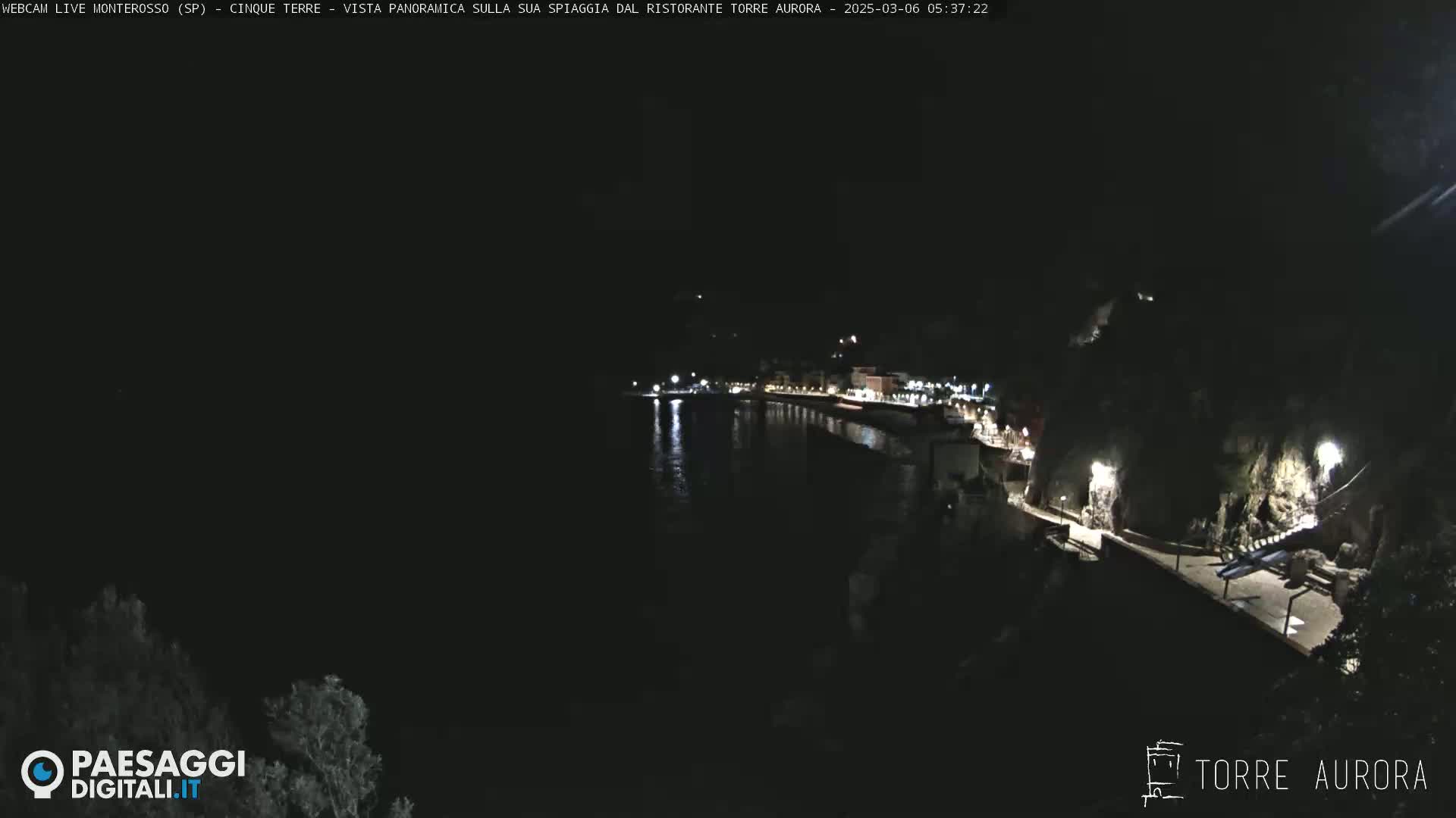 A nighttime, coastal view shows a dark, calm body of water reflecting lights from a shoreline lined with buildings and a walkway.