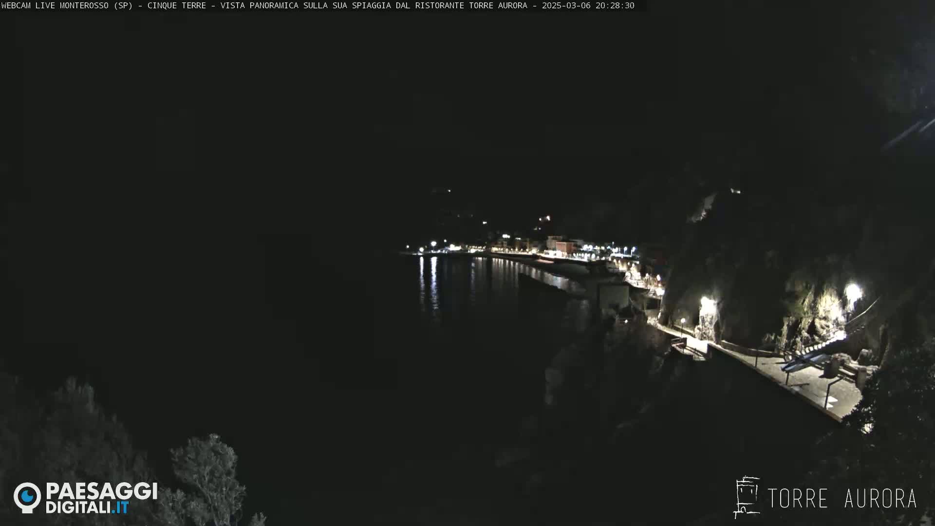 A nighttime, coastal town scene shows a dark, calm body of water reflecting lights from buildings on a cliffside.