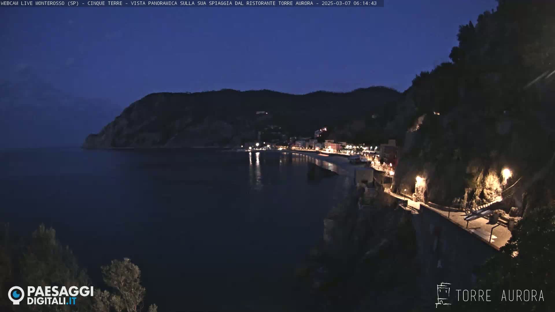 A nighttime coastal town scene shows a dark body of water, illuminated buildings along a shoreline, and steep, dark cliffs under a twilight sky.