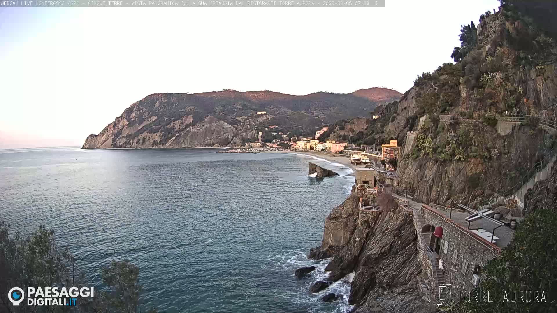 A coastal town nestled between rocky cliffs and a calm sea under a clear, dusky sky.