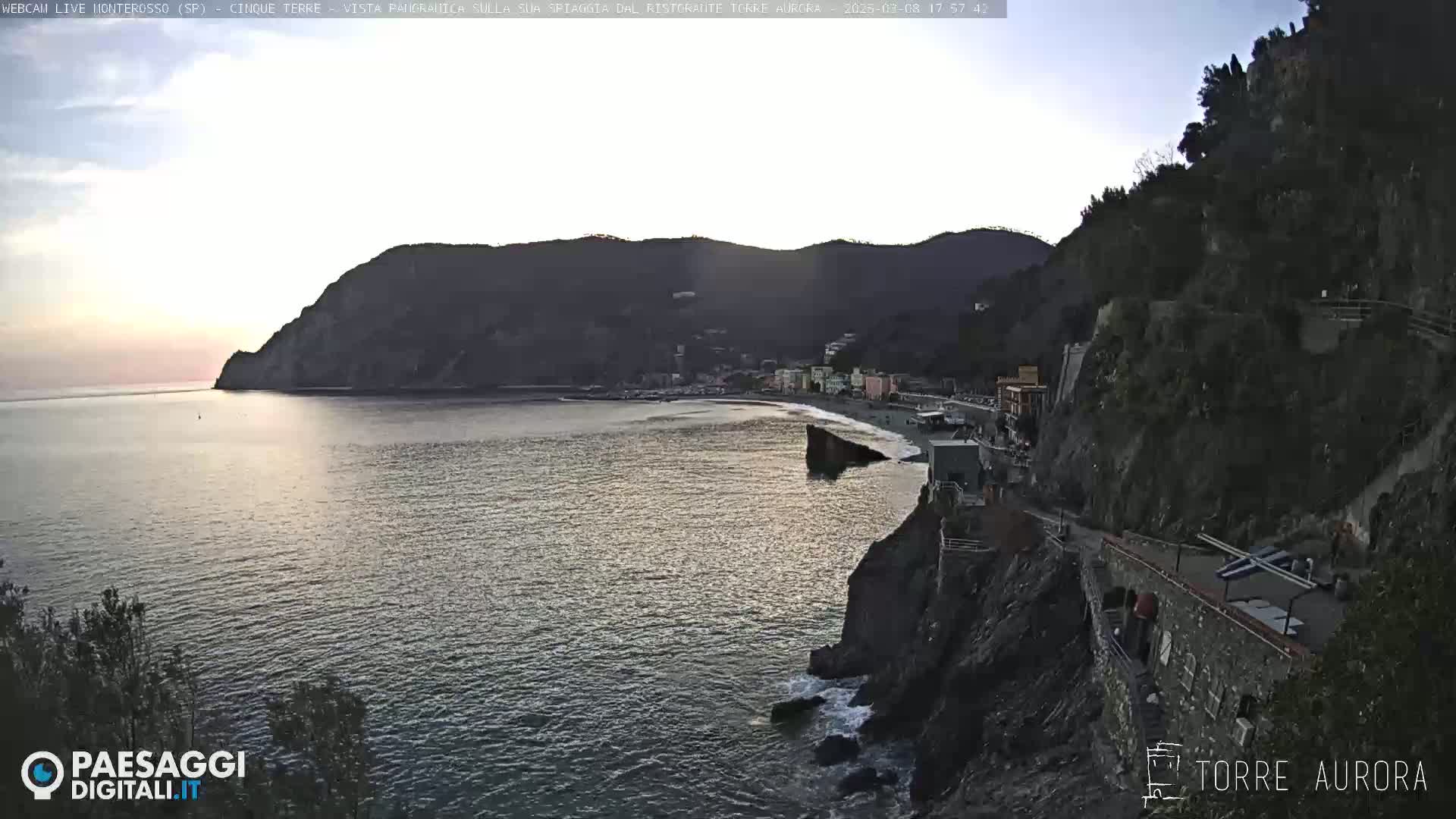 A coastal town nestled between dark hills and a calm sea at sunset.