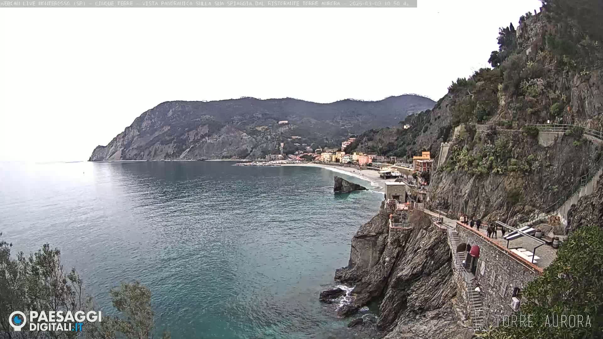 A coastal town nestled between steep, rocky cliffs overlooks a calm, blue sea under an overcast sky.