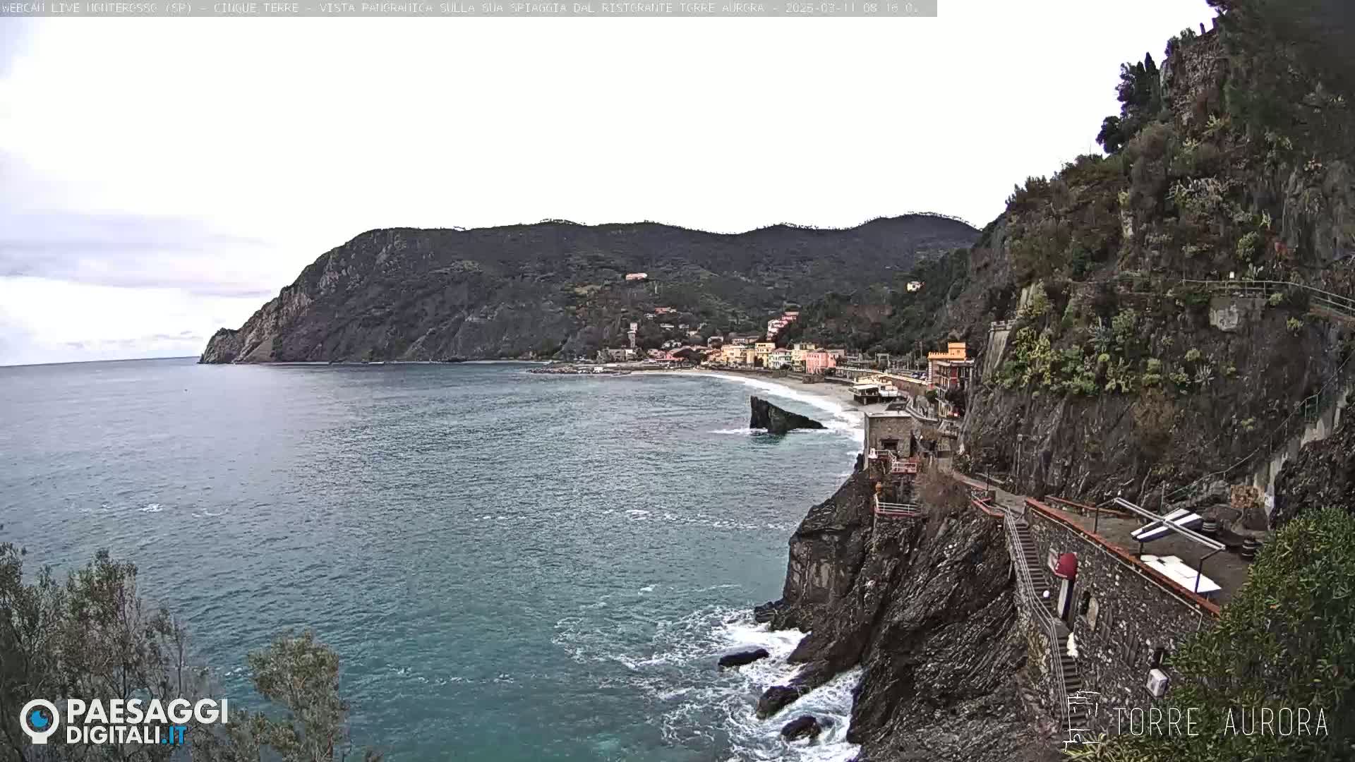 A coastal town nestled between dark, rocky cliffs and a calm sea under an overcast sky.