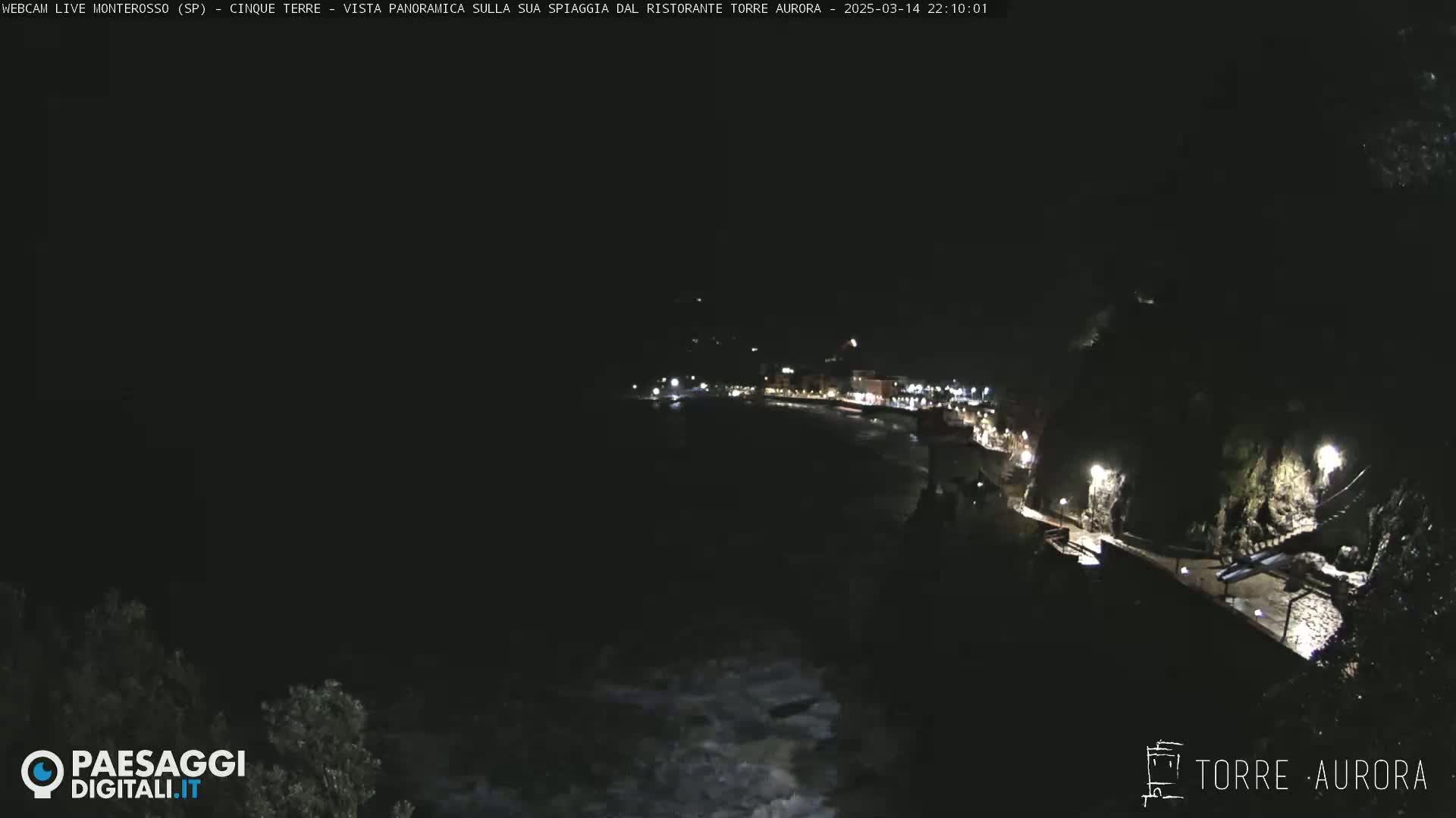 A nighttime, panoramic view of a dark, coastal town with lights illuminating a shoreline path and buildings, partially obscured by darkness.