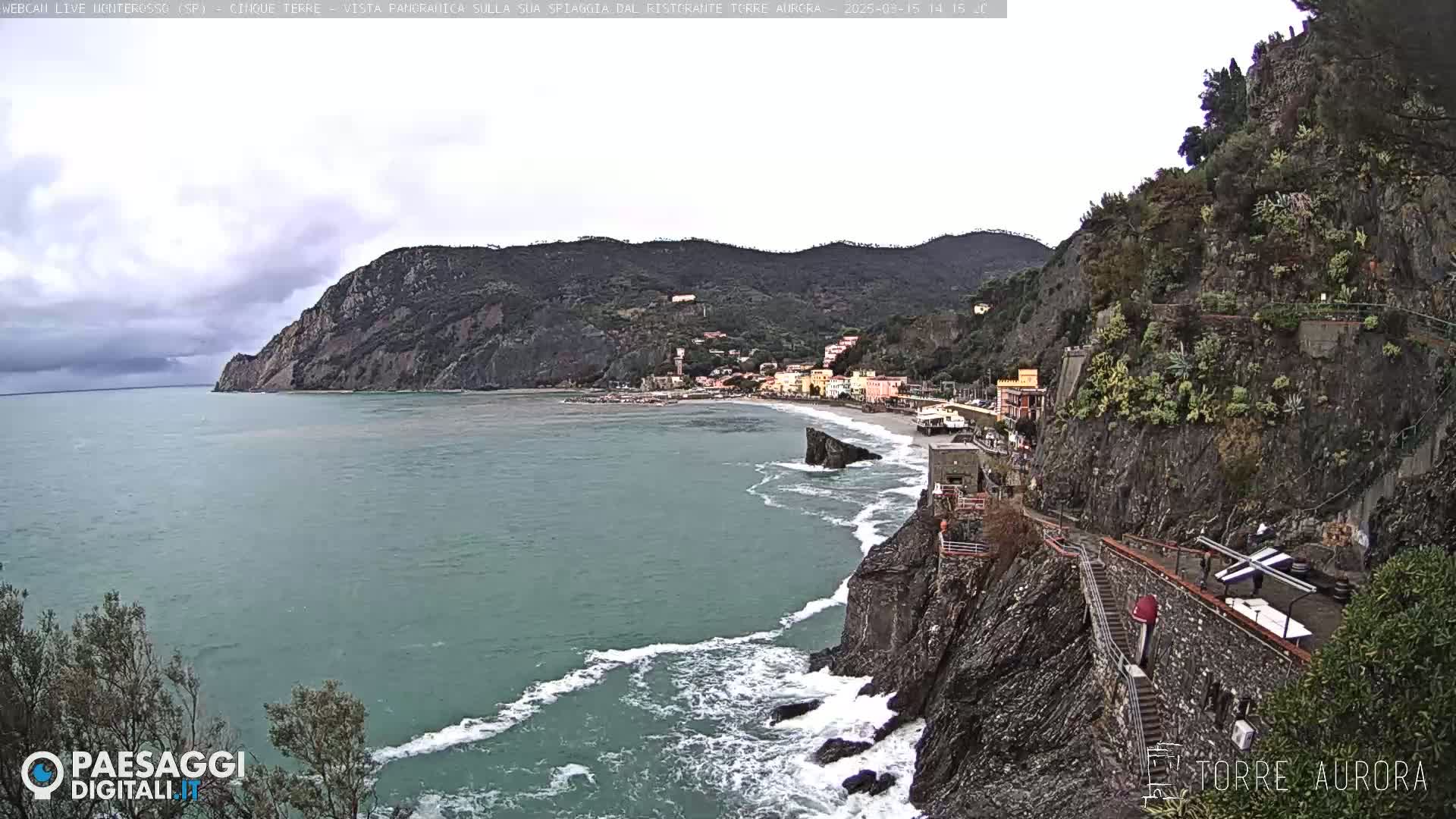 A coastal town nestled between steep cliffs and the sea on an overcast day, with waves crashing against the rocks.