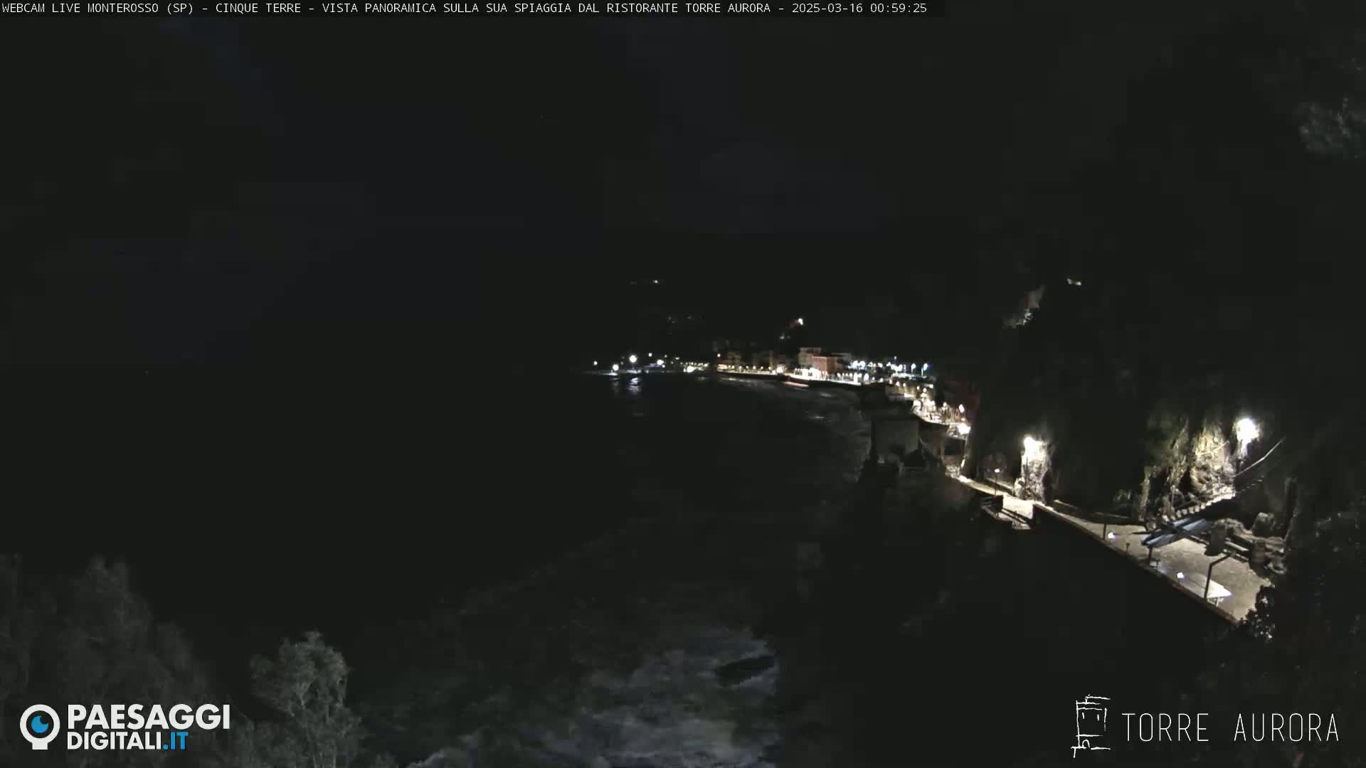 A nighttime, seaside town is shown with a dark, wavy ocean and dimly lit buildings along a paved walkway.