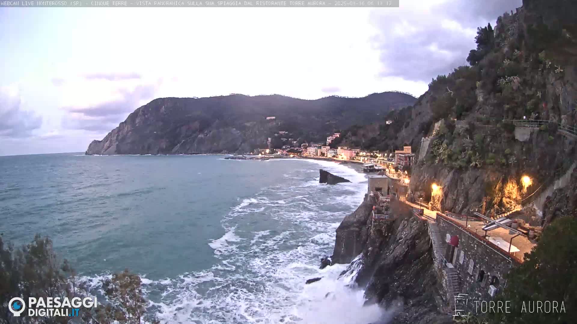 A coastal town nestled between a rocky cliff and the sea, with moderate waves crashing against the shore under an overcast sky.