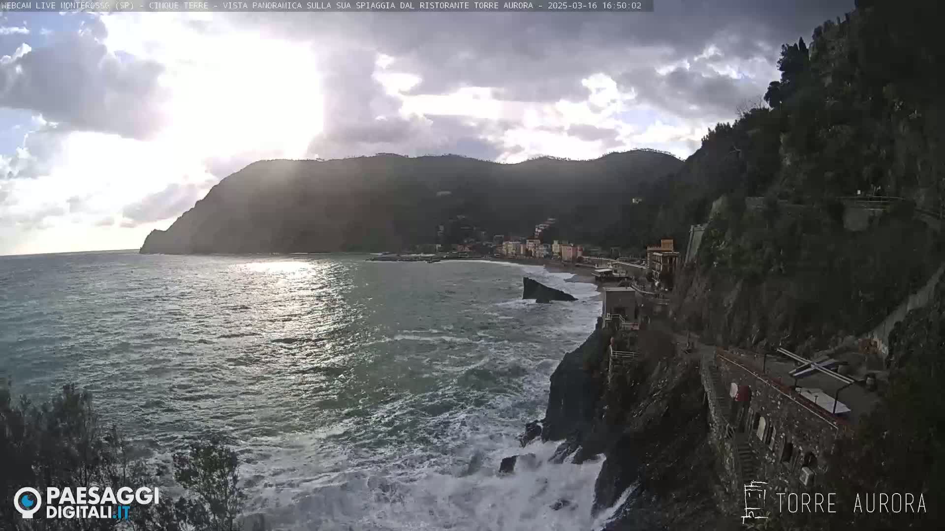 A seaside town nestled between dark, rocky cliffs is partially obscured by overcast skies, with waves crashing against the shoreline.