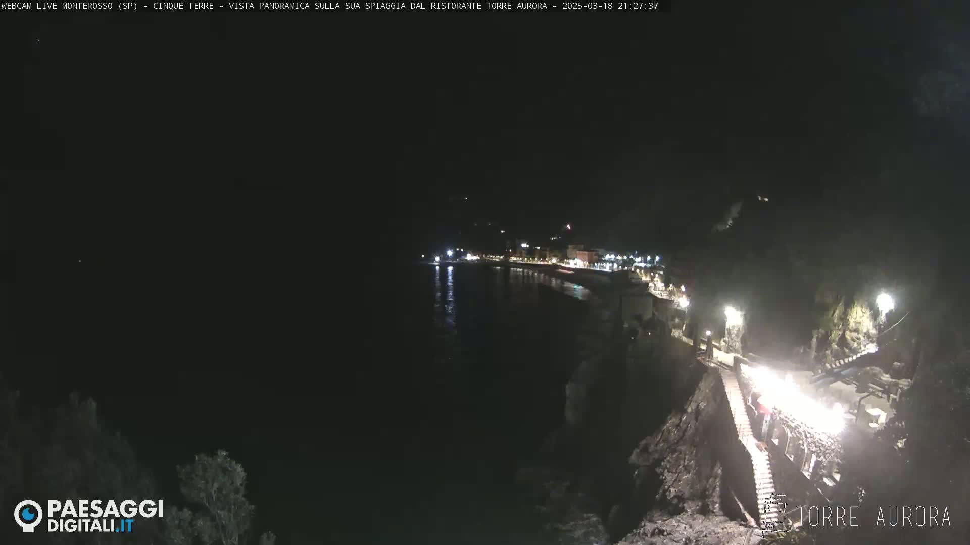A nighttime coastal scene shows a dark body of water reflecting lights from a town along the shore, with stairs leading down a rocky cliff toward the water's edge.