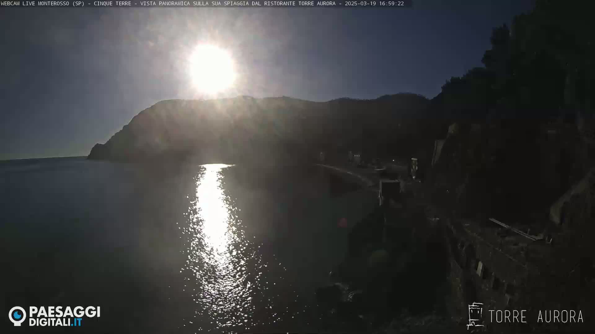 A bright sun shines over a calm body of water, reflecting off the surface near a dark, rocky shoreline and hillside.