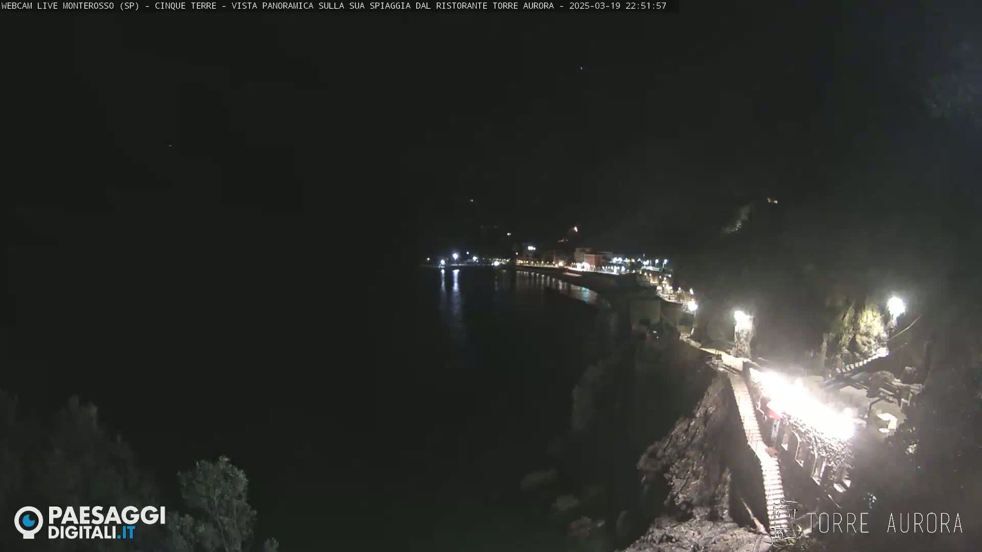A nighttime, panoramic view shows a dark coastline with a village's lights reflecting on the water, and stairs leading down to a rocky shore.