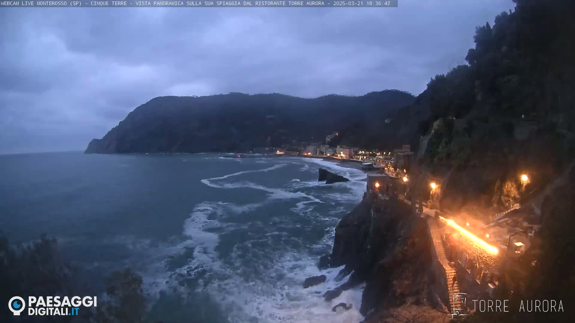 A rocky coastline at dusk, with a town nestled against the cliffs and rough waves crashing against the shore under a cloudy sky.