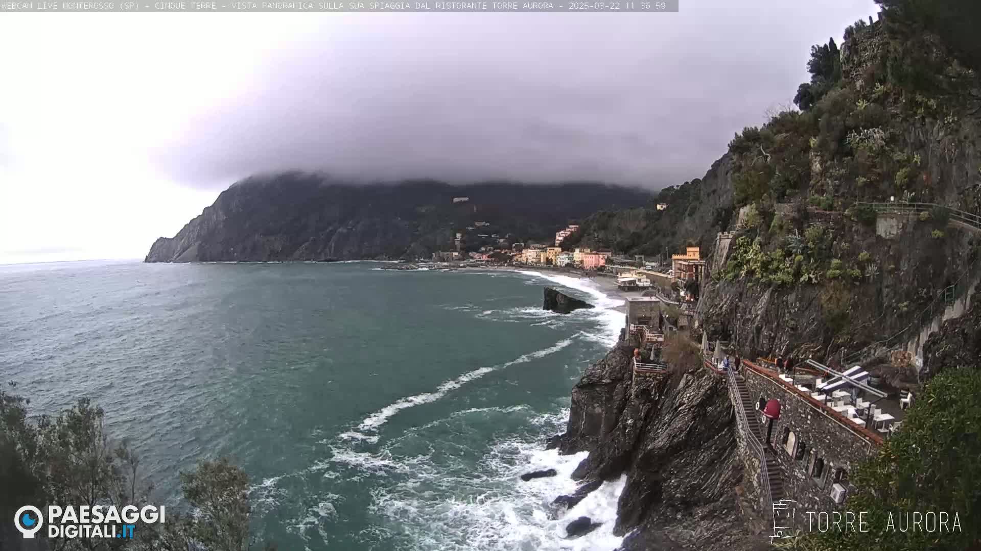 A coastal town nestled between rocky cliffs, partially obscured by fog, is seen from an elevated vantage point overlooking a turbulent sea.