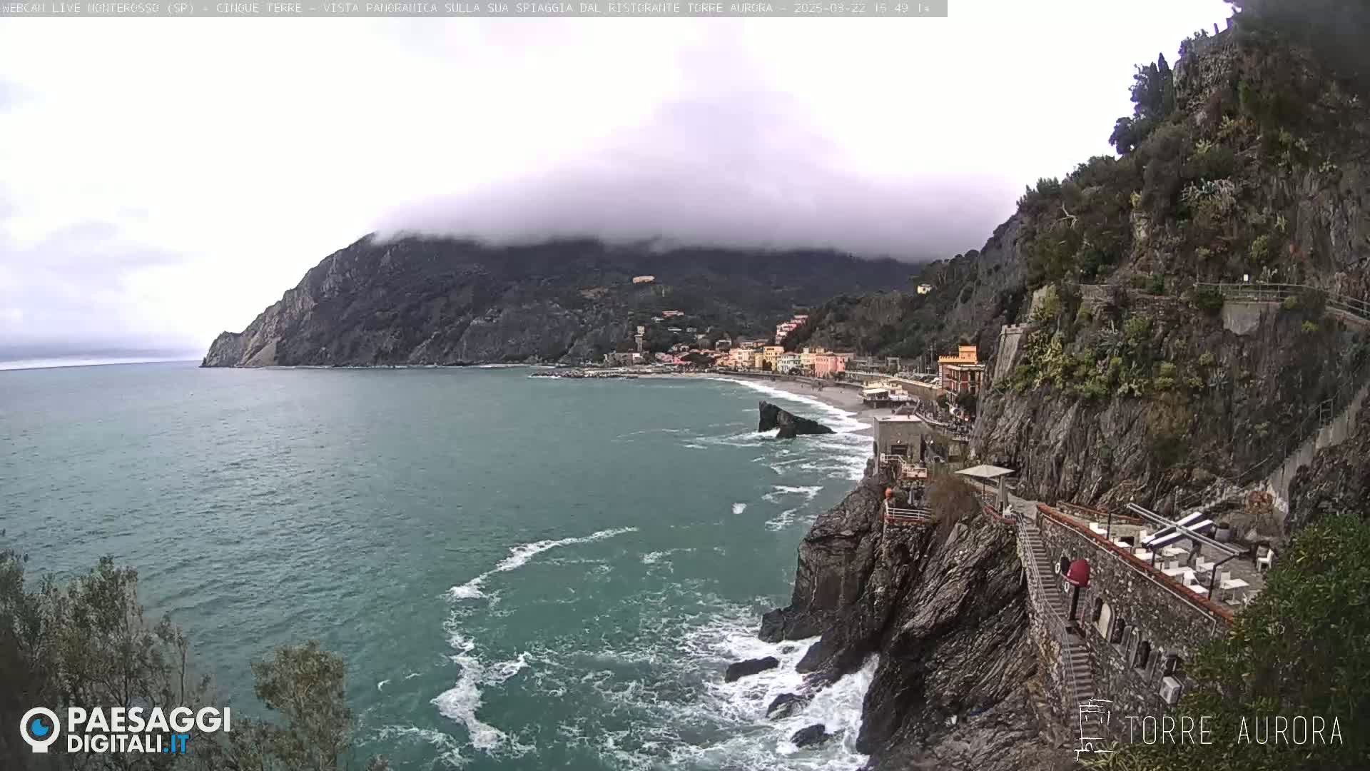 A coastal town nestled between a rocky cliff and the sea, partially obscured by low-hanging clouds.