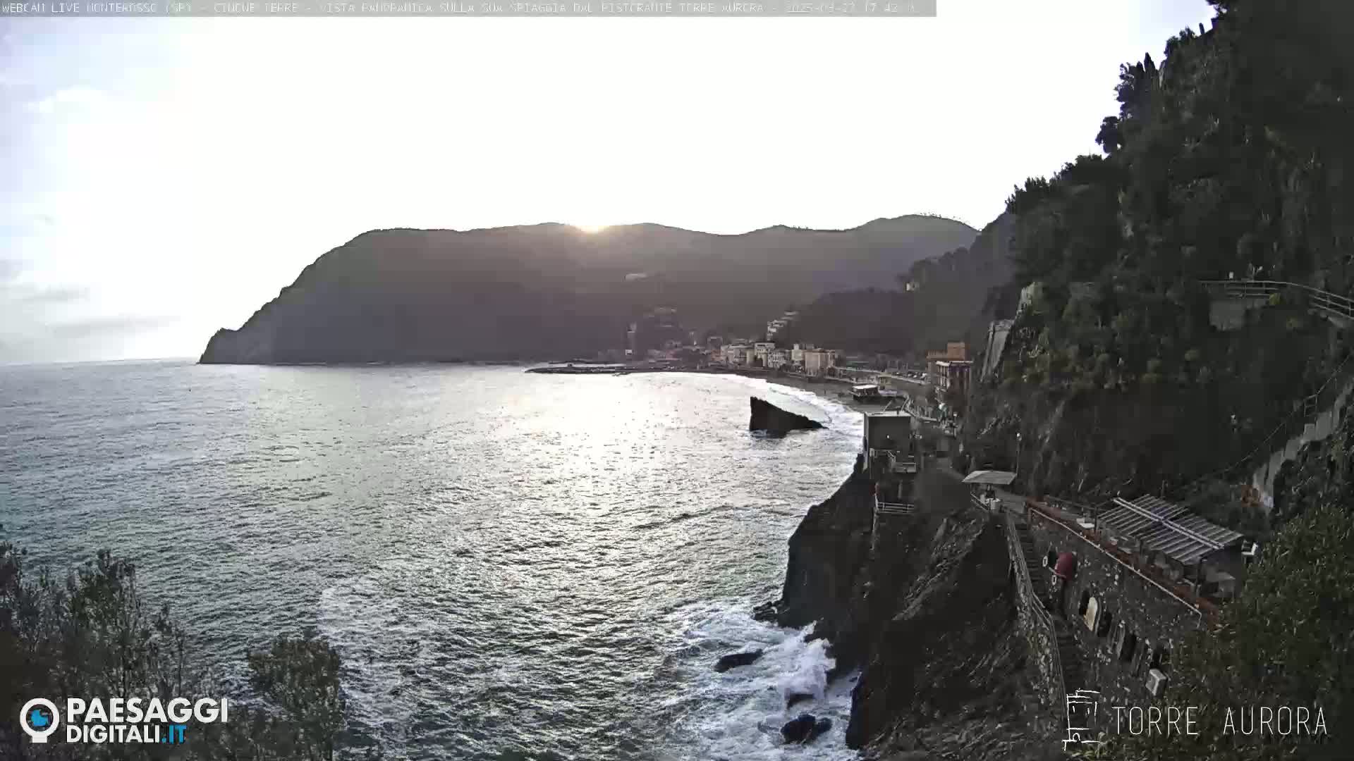 A coastal town nestled between a steep cliff and the sea under a partly cloudy sky, with waves crashing against the rocks below.