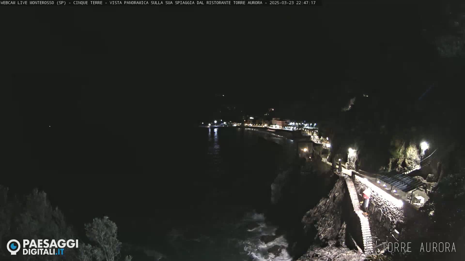 A nighttime coastal scene shows a dark body of water with waves lapping against a rocky shoreline, where a path with lights leads to a building illuminated against a dark, starless sky.