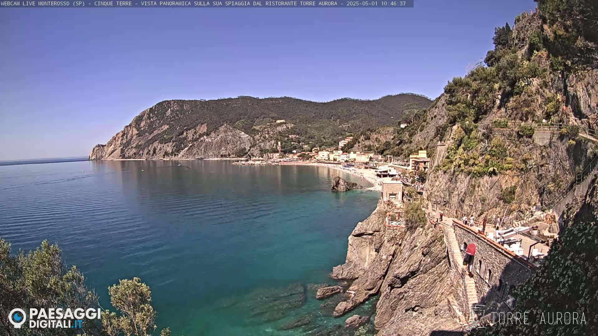 A panoramic view shows a clear, blue ocean meeting a rocky coastline with a town and sandy beach nestled into the hillside under a bright, sunny sky.