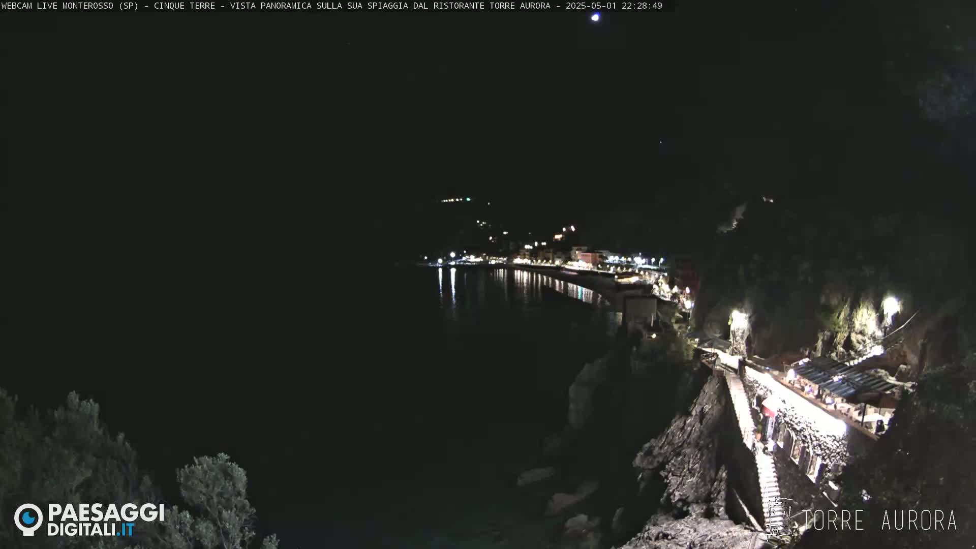 A nighttime, panoramic view of a coastline with a village’s lights reflecting on dark water, seen from a high vantage point overlooking a seaside restaurant.