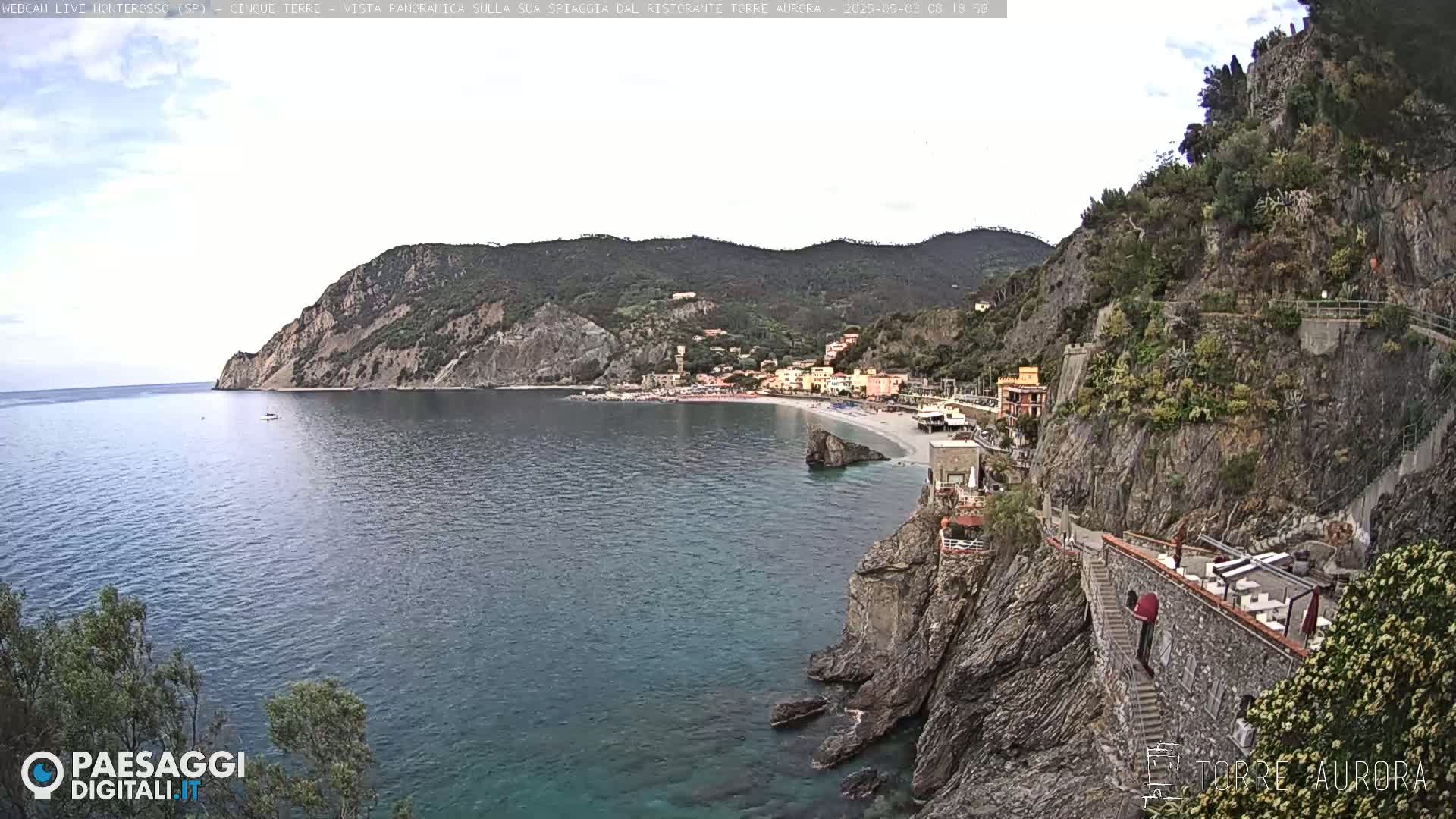 A coastal village with a sandy beach is nestled between rocky cliffs, overlooking a calm sea under a mostly cloudy sky.