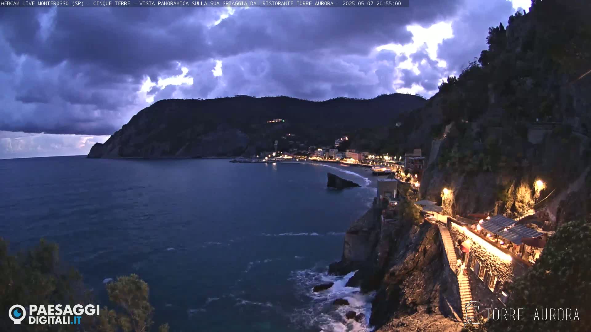 A nighttime coastal scene shows a town nestled between dark hills and a dark-blue sea under a partly cloudy sky.