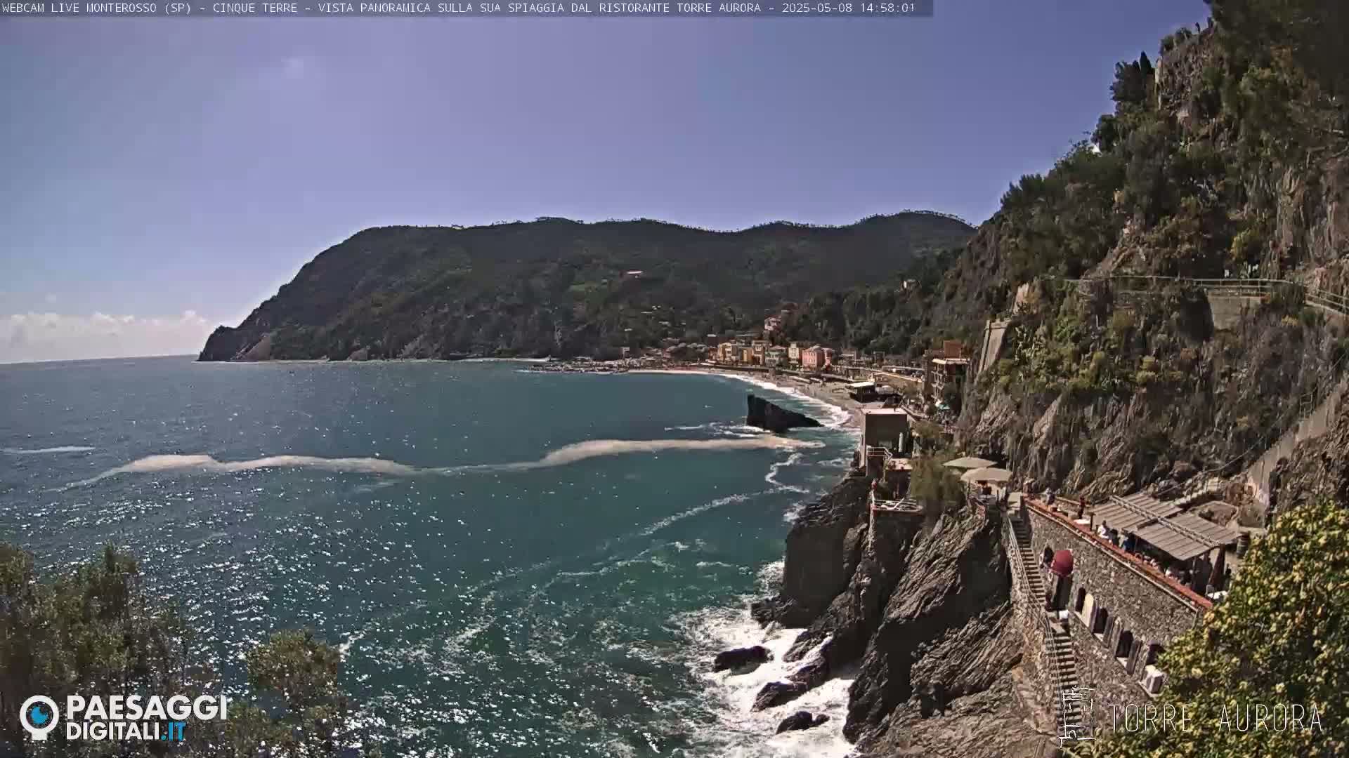 A sunny coastal scene shows a rocky cliffside with buildings and a beach below, overlooking a dark blue sea with small waves.