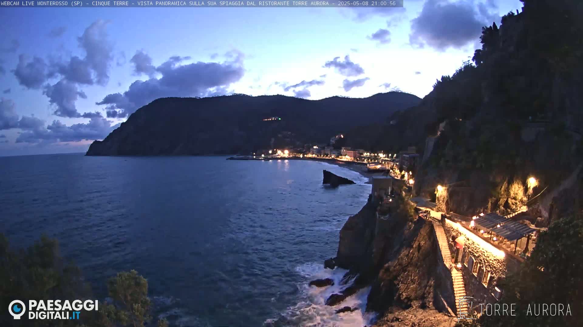 A coastal town at twilight is nestled between dark hills and a calm, dark-blue sea, with waves lapping against rocky shores.