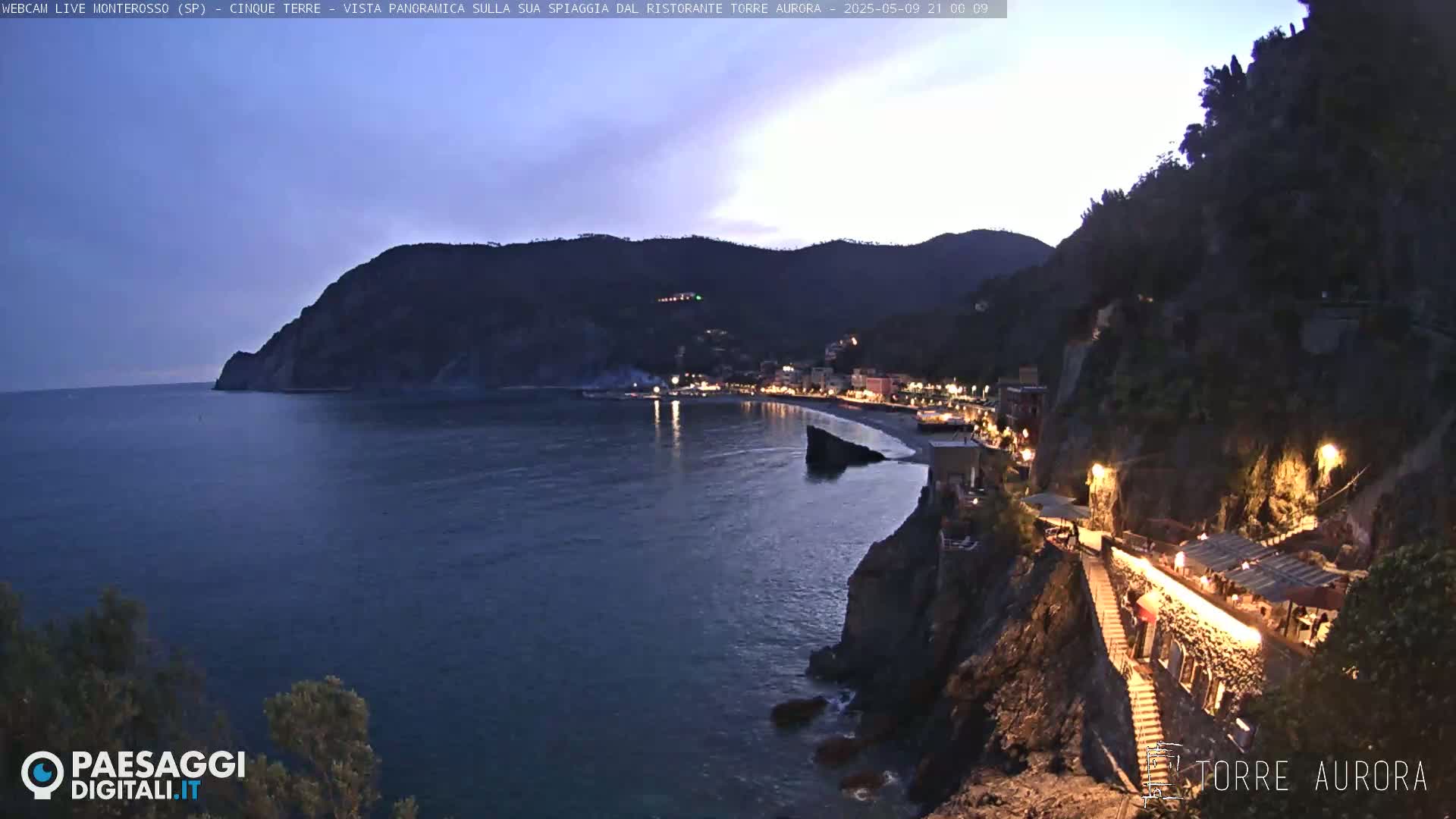 A coastal town at twilight is nestled between dark hills and a calm, dark-blue sea.