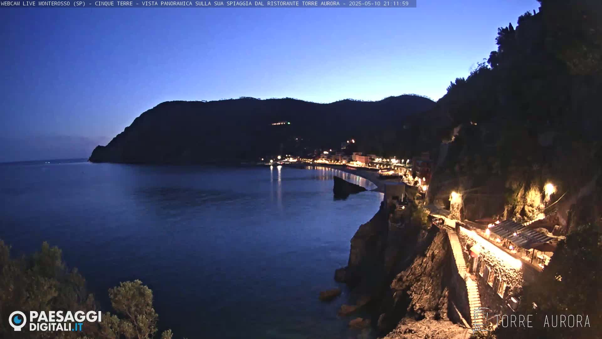 A nighttime, panoramic view of a coastal town nestled between a dark mountain and a calm, dark-blue sea, illuminated by lights.
