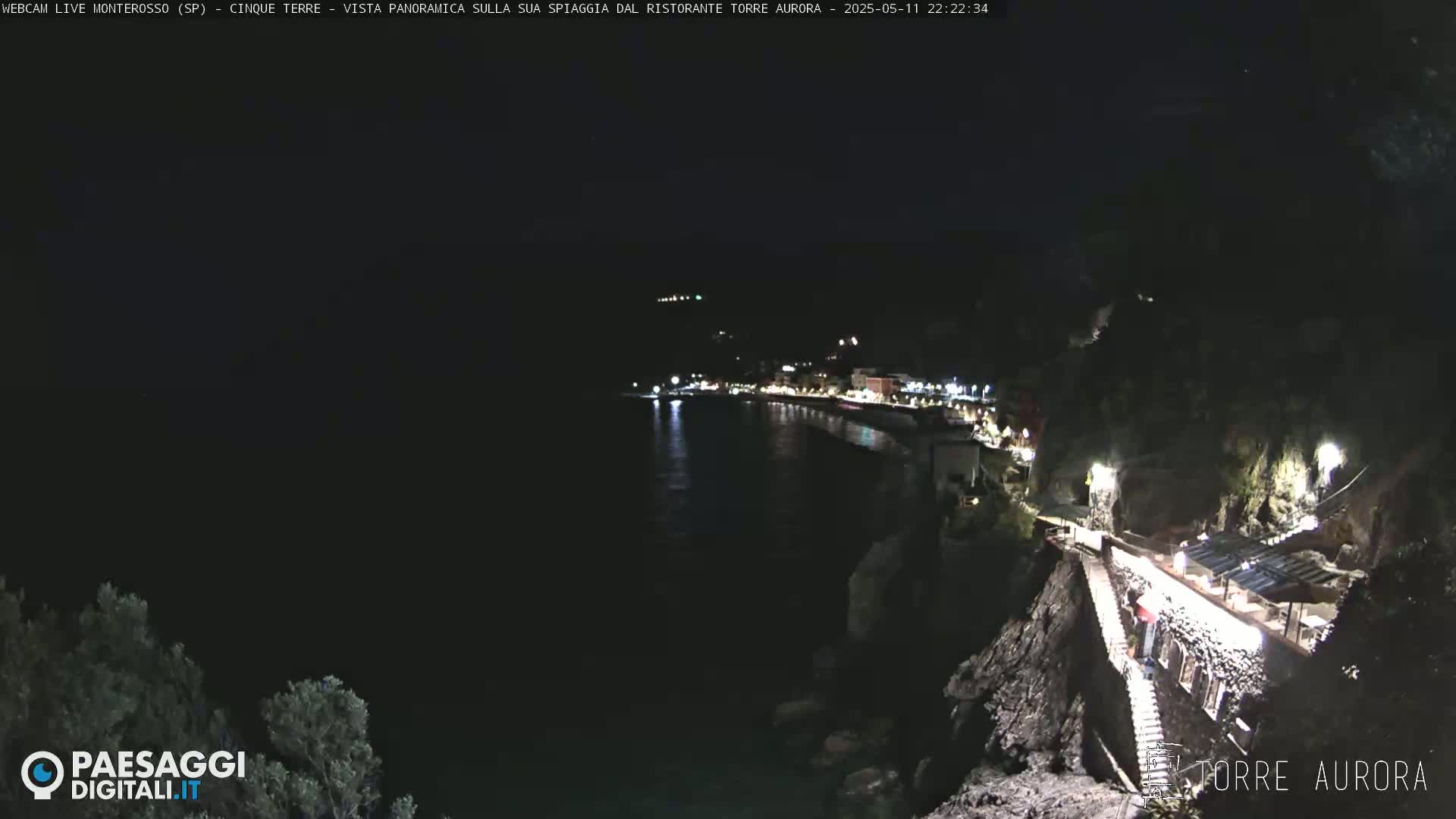 A nighttime, coastal view shows a dark body of water with a dimly lit town on the opposite shore, and a stone building with lights on a cliff to the right.