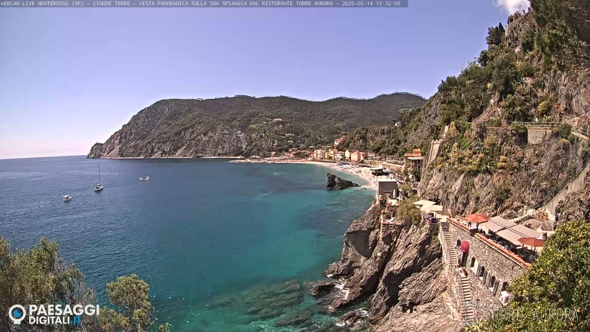 A clear, sunny day overlooks a coastal town nestled between rocky cliffs and a calm, turquoise sea with several small boats visible.