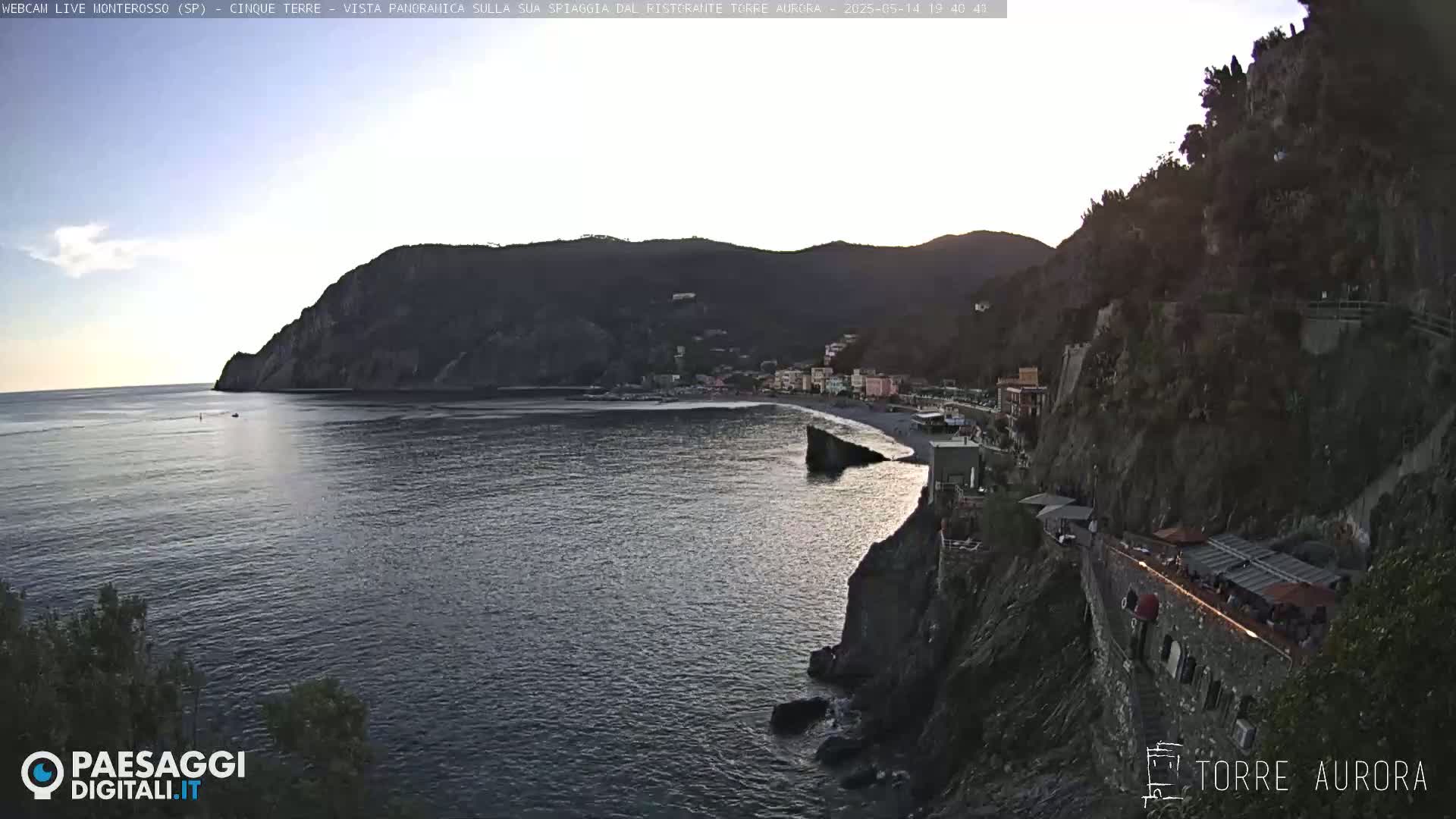 A coastal town nestled between dark cliffs and a calm sea is bathed in the soft light of sunset.