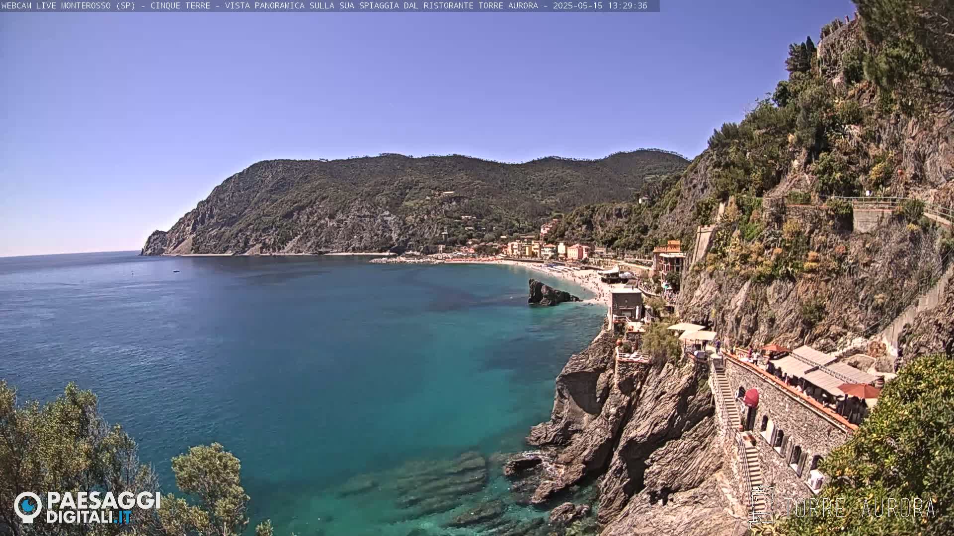 A clear, sunny day reveals a coastal town nestled between rocky cliffs and a calm, teal sea.